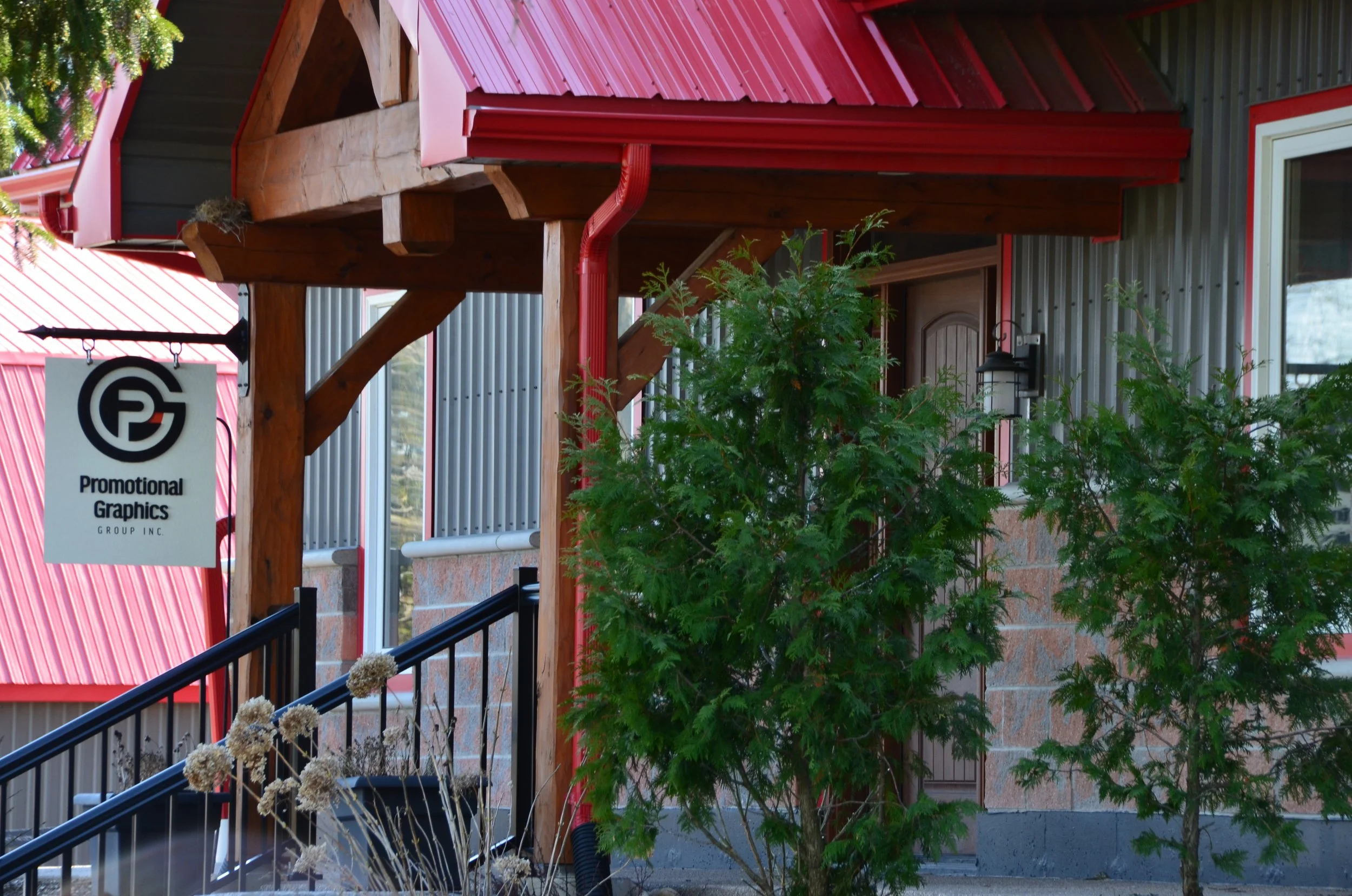 Exterior of a building with a sign that reads 'Promotional Graphics Group Inc.', a front porch with a black railing, green trees, and a red metal roof.
