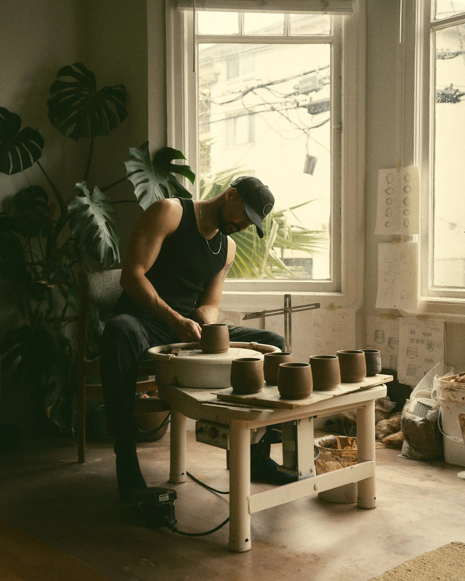 Dan Mack using a pottery wheel indoors near a window with several clay pots and plants visible.