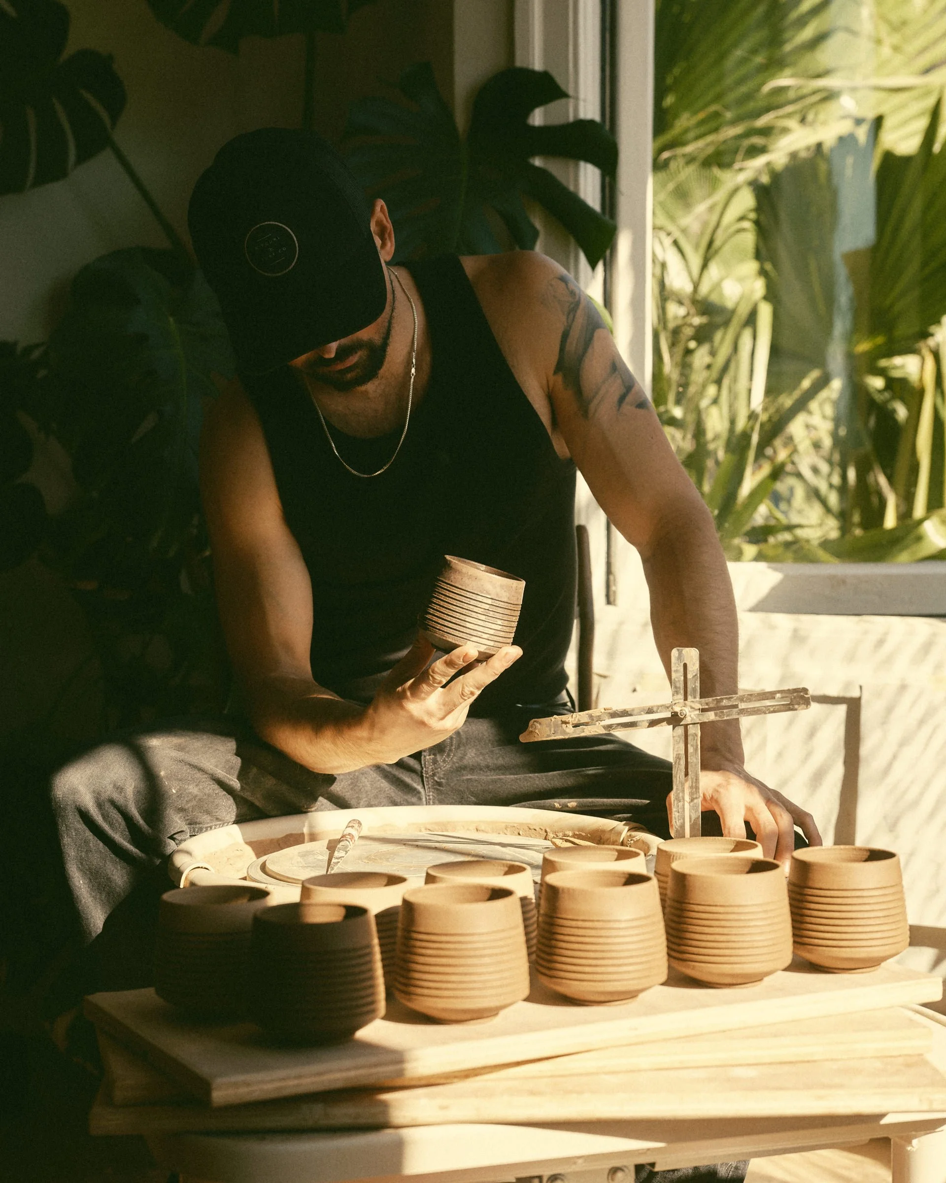 Dan mack in his studio in black tank top and cap making pottery, sitting at a pottery wheel surrounded by ceramic cups, with tropical plants in the background. 