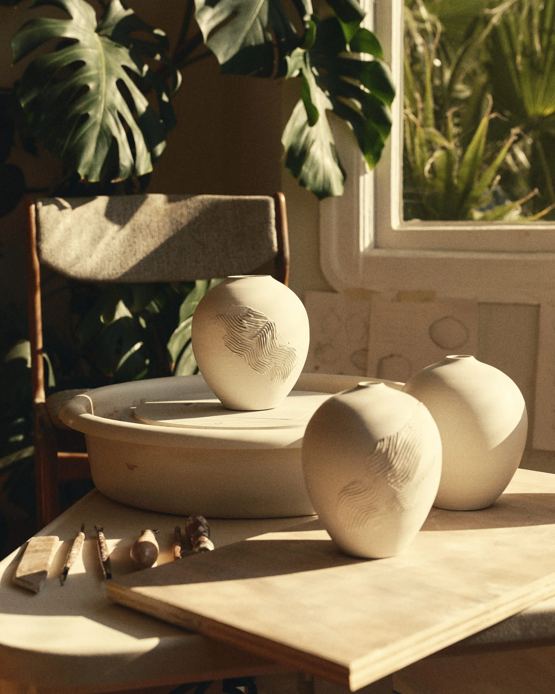 Ceramic vases with carved patterns on a table, surrounded by pottery tools, with a window and green plants in the background.