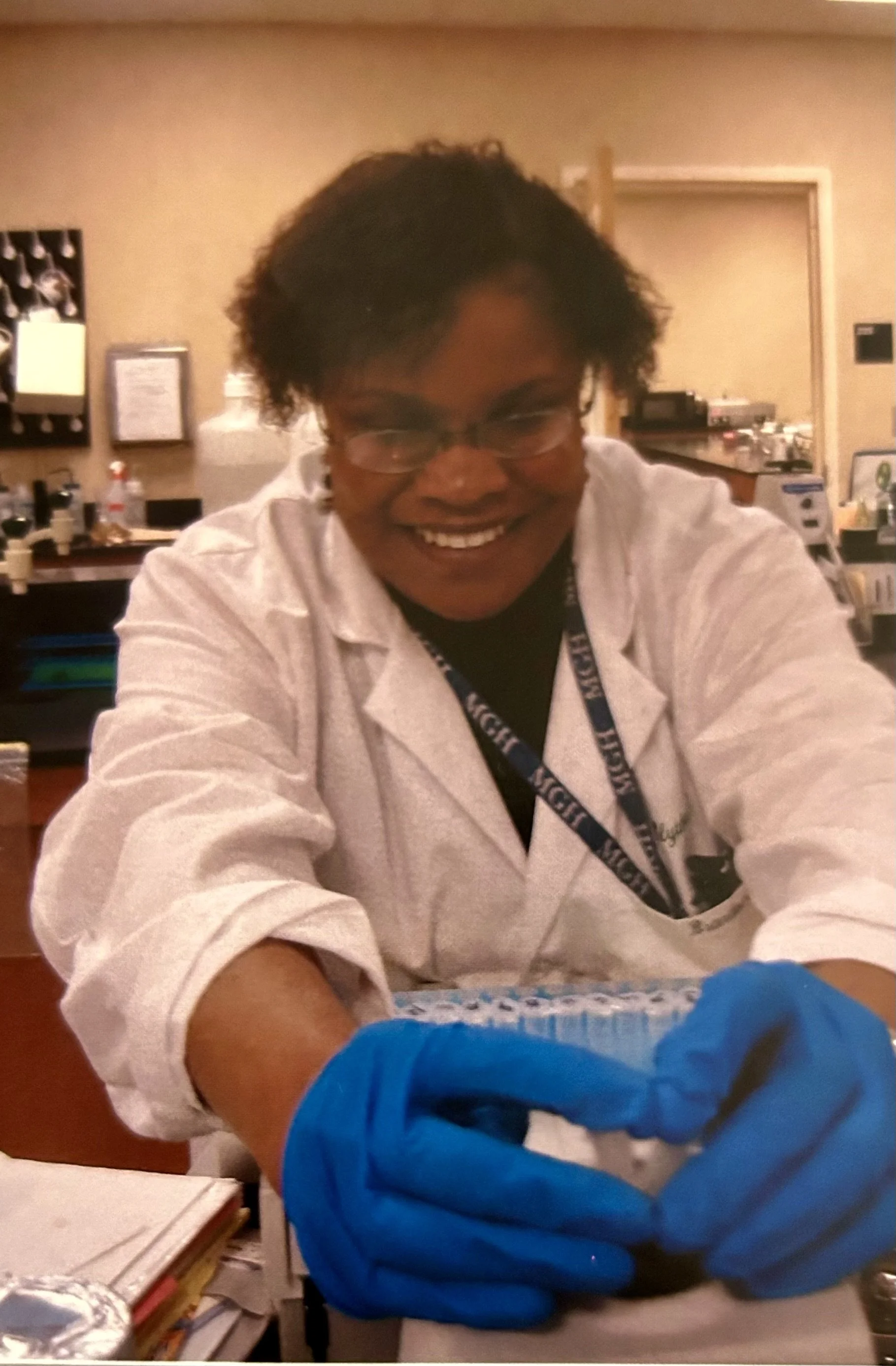A woman in a white lab coat and blue gloves smiling, working in a laboratory setting.