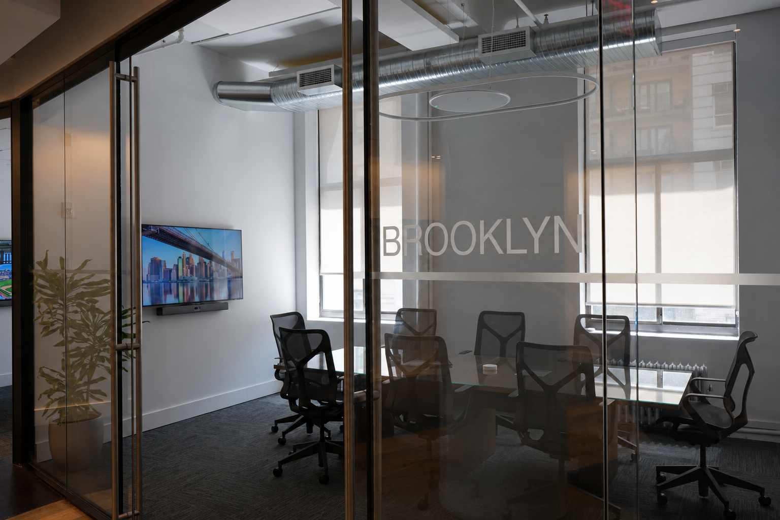Modern conference room with a long table, brown chairs, digital screen, and windows on the right.