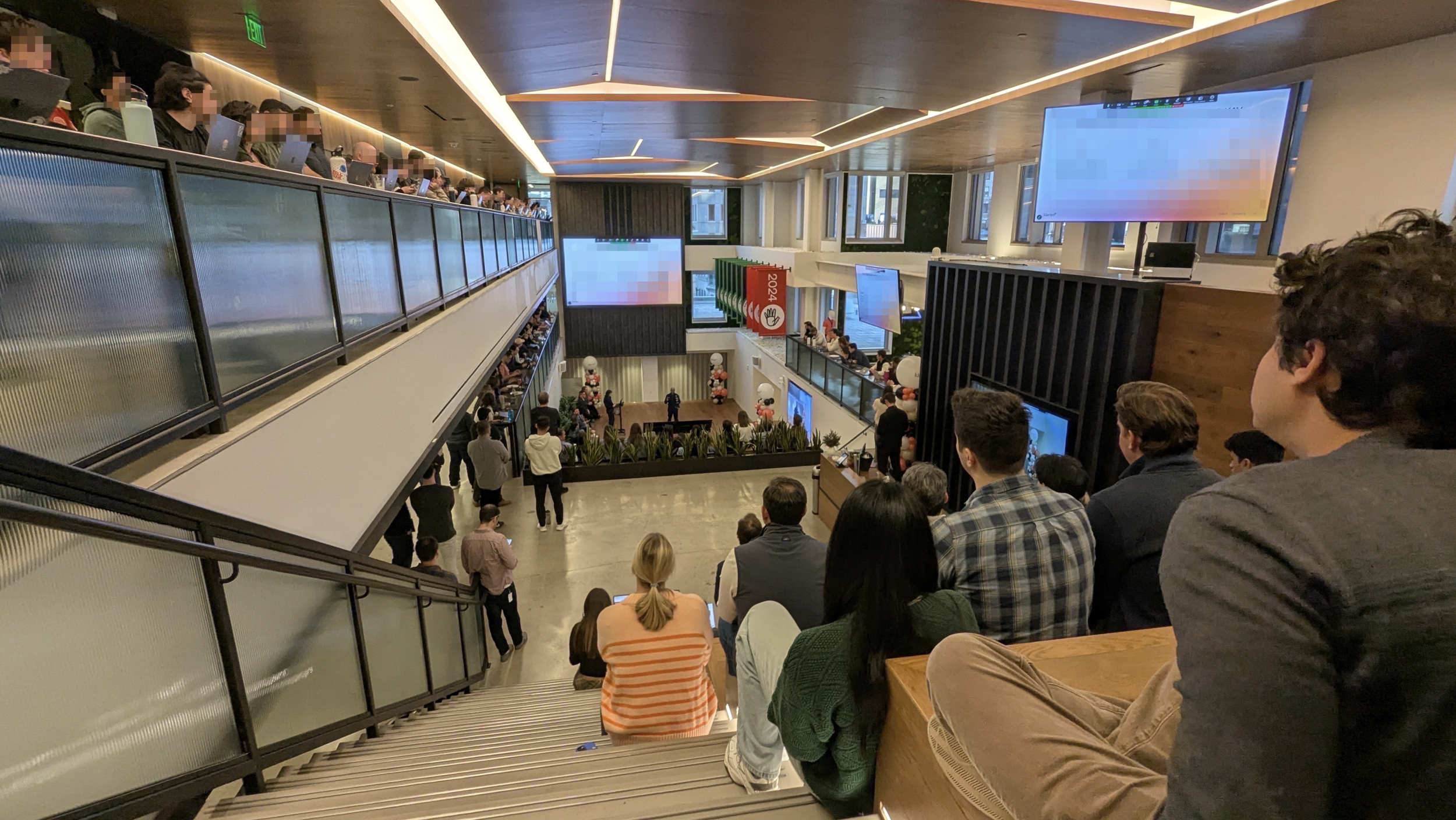 People gathered in a modern, multi-level indoor space with large screens, attending a presentation or meeting.
