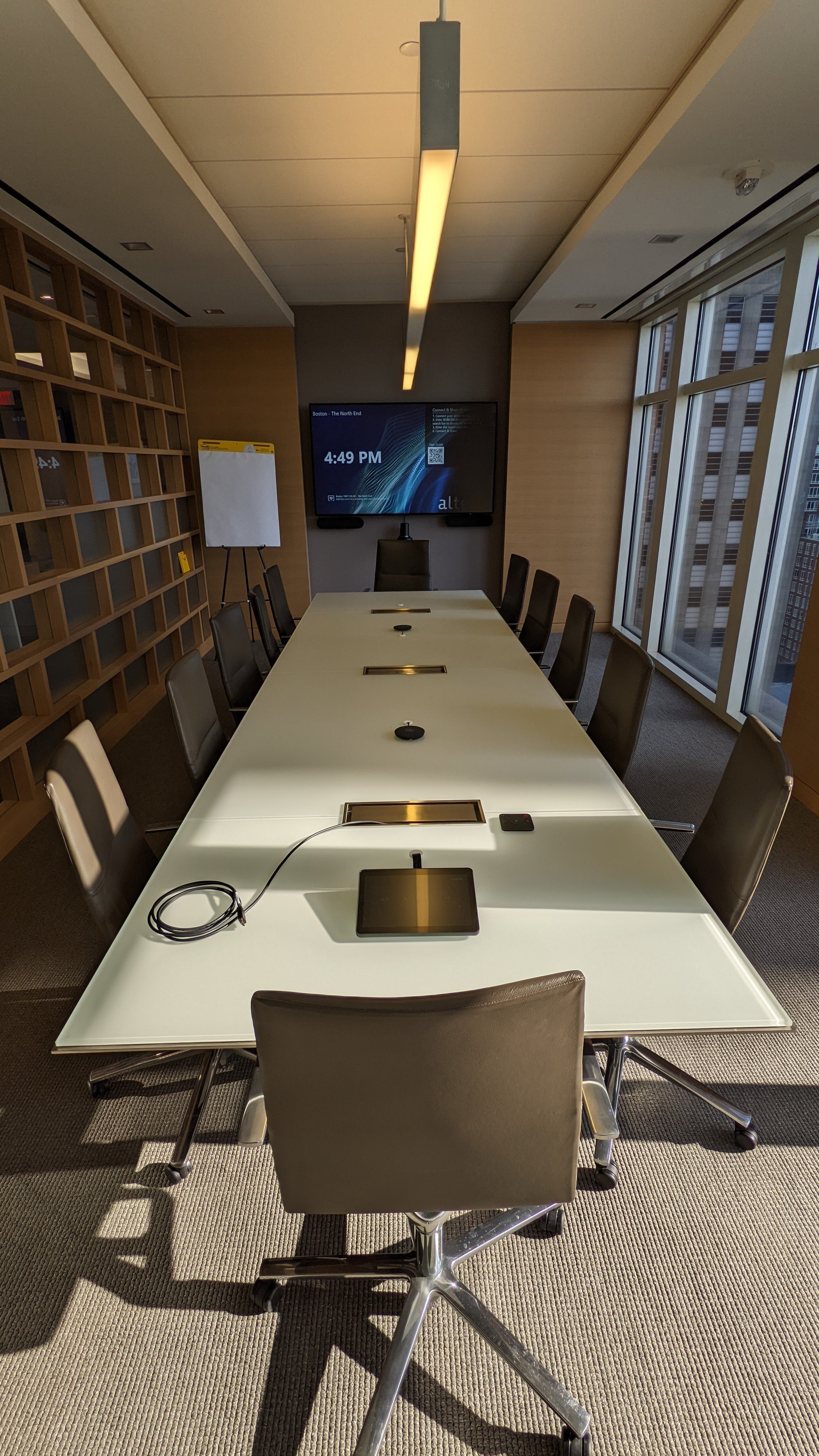 Modern conference room with a long table, brown chairs, digital screen, and windows on the right.