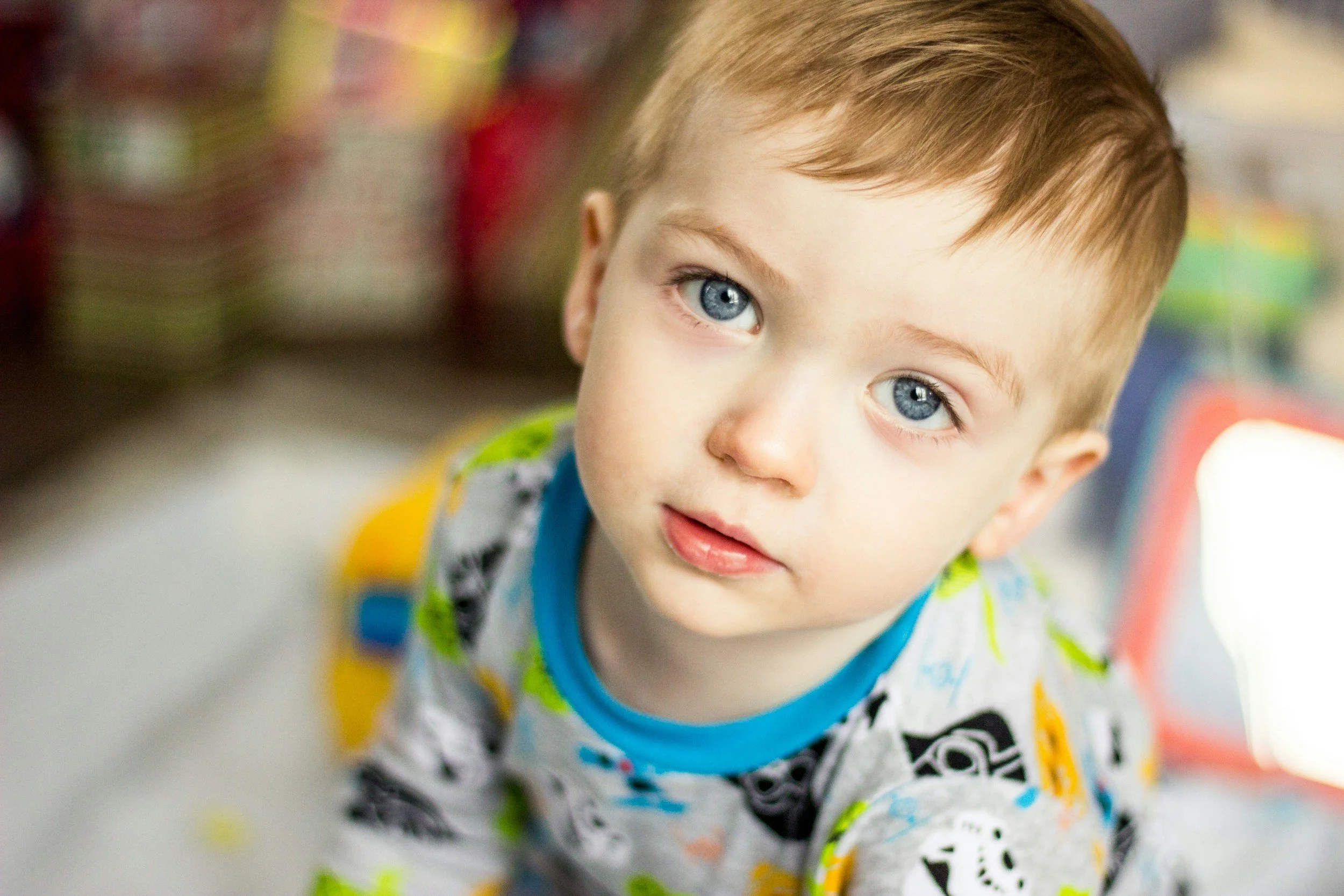 Close-up of a young boy with blue eyes and light brown hair, wearing a colorful patterned shirt, looking up at the camera indoors.