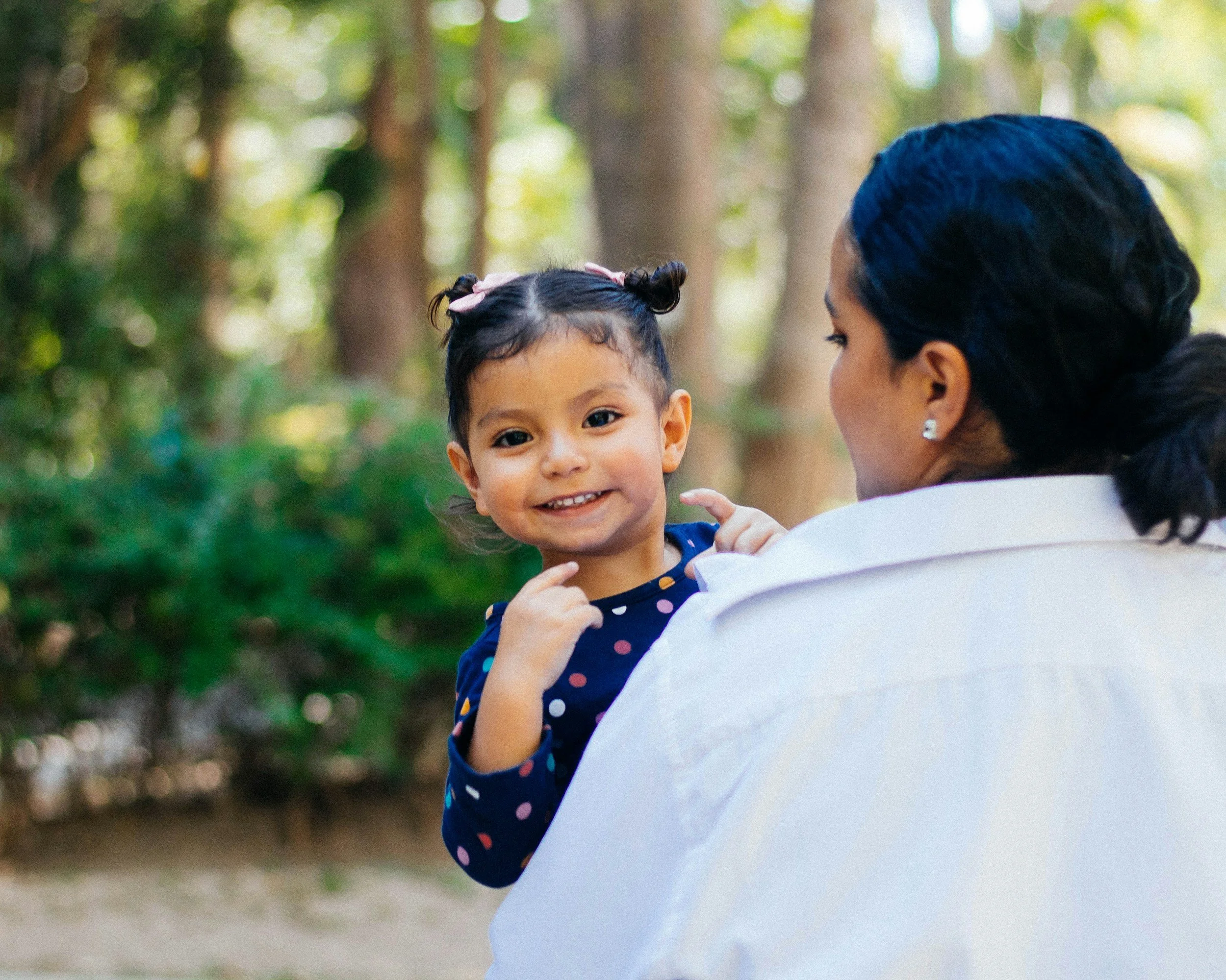 A young girl with dark hair styled in two buns, smiling and looking at the camera, held by a woman with dark hair pulled back, outdoors in a wooded area.