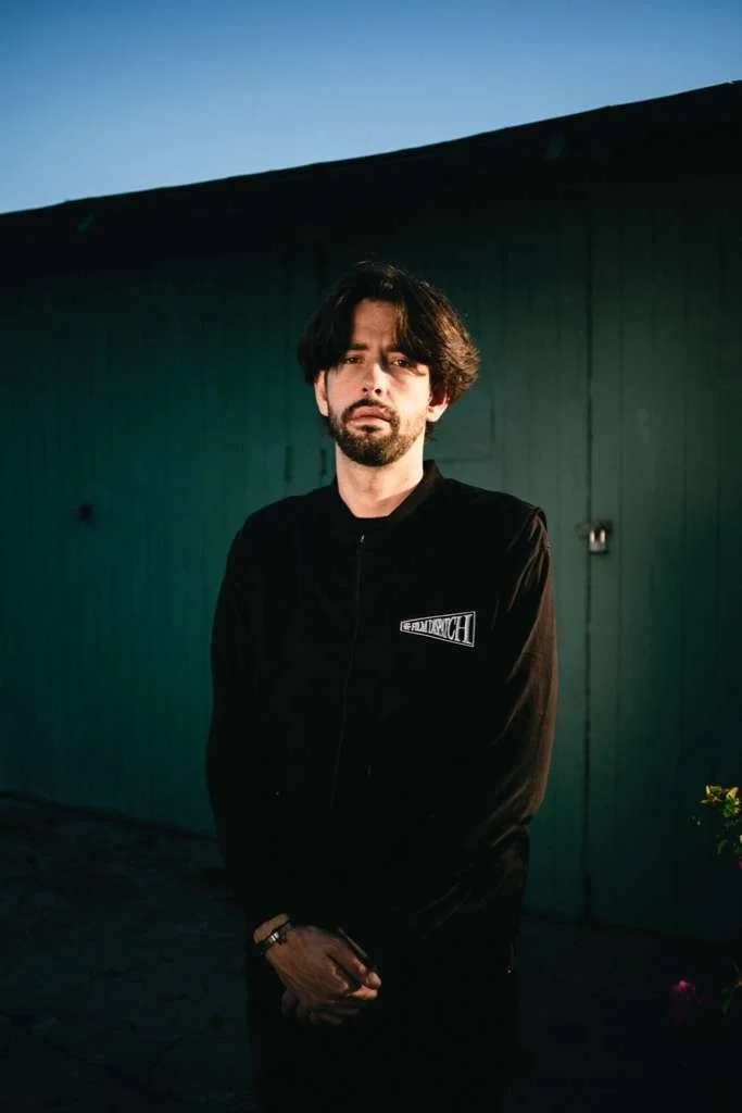 A young man with dark, wavy hair and a beard standing outdoors in the evening, wearing a black jacket with a Patchy logo, in front of a green wooden wall.