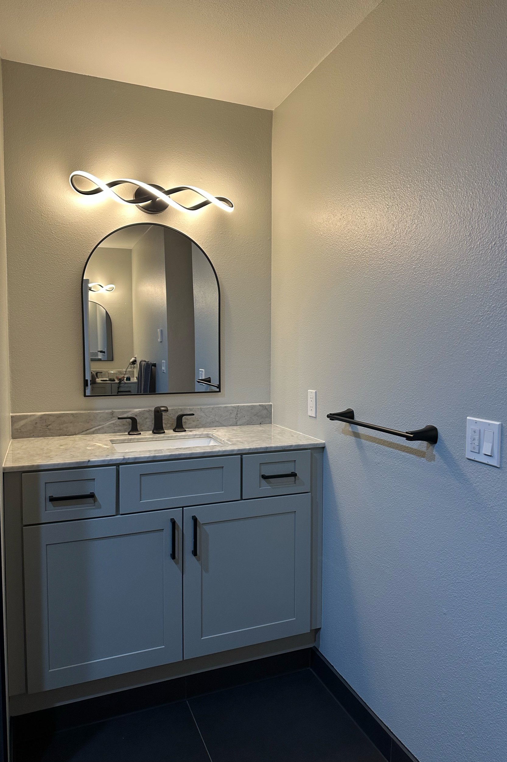Modern bathroom vanity with gray cabinet, marble countertop, and black hardware; features an artistic mirror and looped light fixture above.