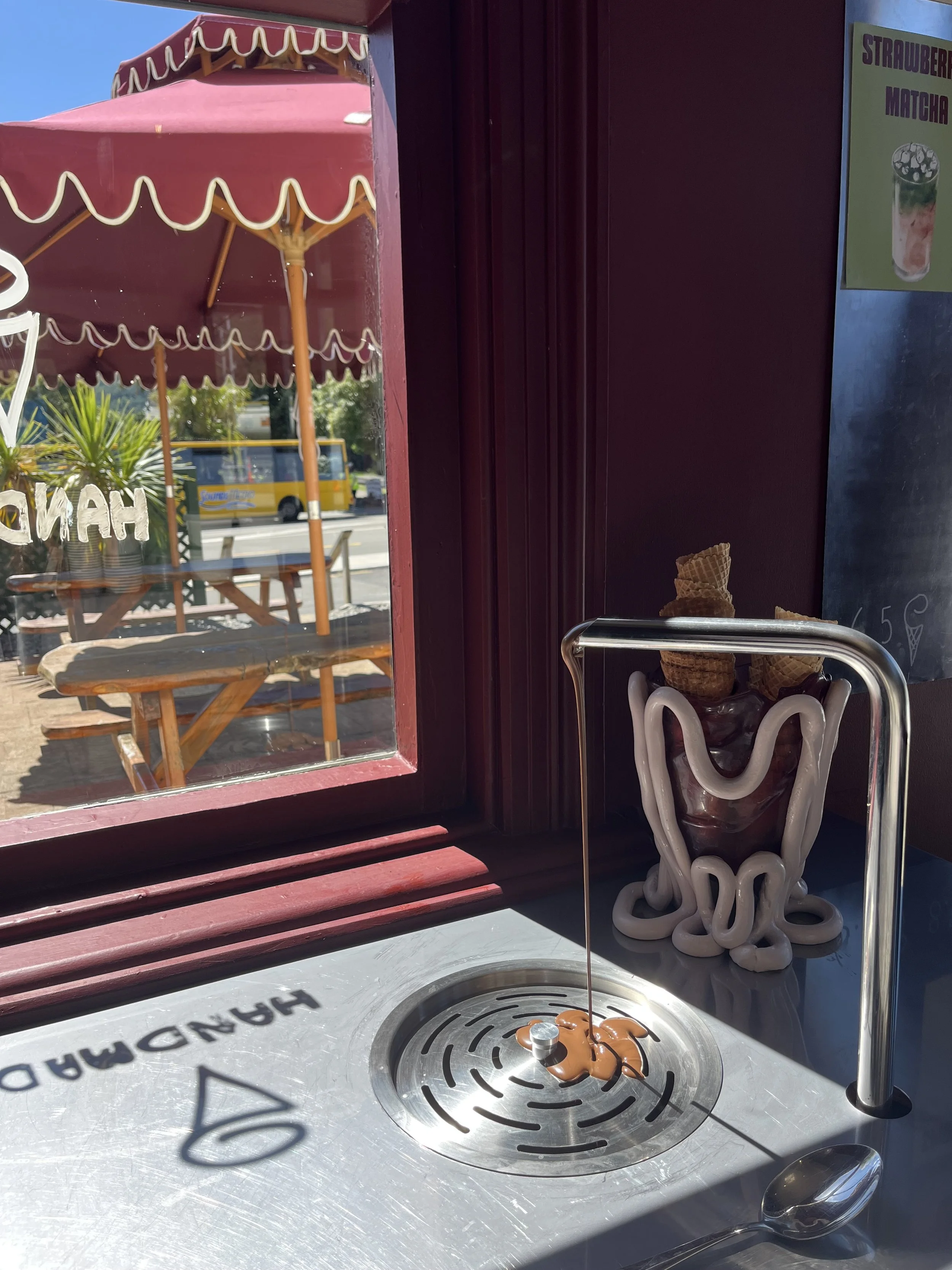 Indoor view of an ice cream shop counter with a swirl of chocolate and vanilla ice cream on a metal surface, with a painted chocolate syrup spill in an oval shape, next to a small deflated balloon with a string, a spoon, and a decorative container holding waffle cones. Outside through a window, there is a pink umbrella with white trim, a picnic table, some greenery, and a yellow bus on the street.