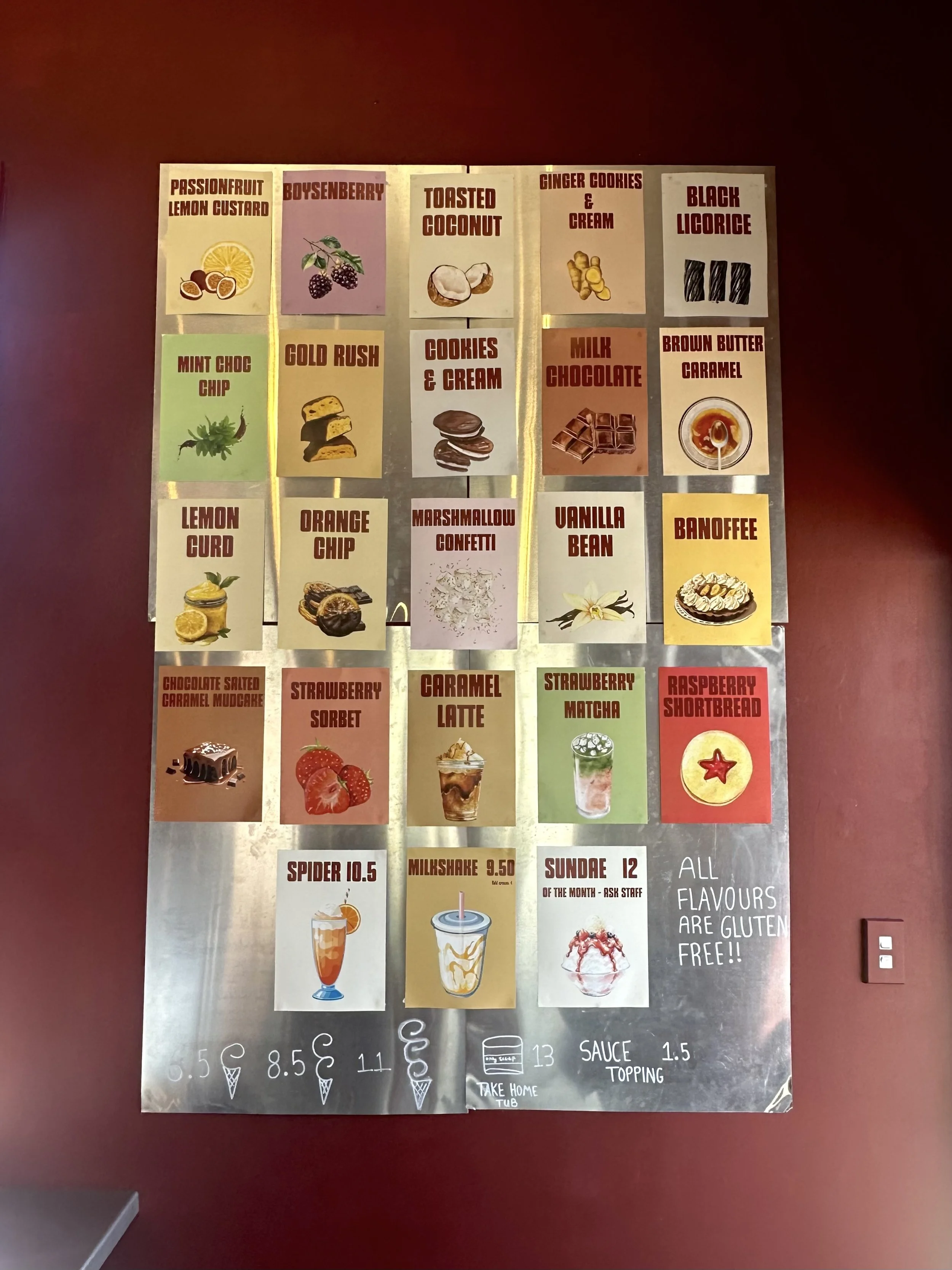 Menu board with various ice cream flavors and toppings, including Passionfruit Lemon Custard, Boysenberry, Toasted Coconut, Ginger Cookies & Cream, Black Licorice, Mint Choc Chip, Gold Rush, Cookies & Cream, Milk Chocolate, Brown Butter Caramel, Lemon Curd, Orange Chip, Marshmallow Confetti, Vanilla Bean, Banoffee, Chocolate Salted Caramel Mudcake, Strawberry Sorbet, Caramel Latte, Strawberry Matcha, Raspberry Shortbread, Spider, Milkshake, Sundae, with prices and notes that all flavors are gluten-free.