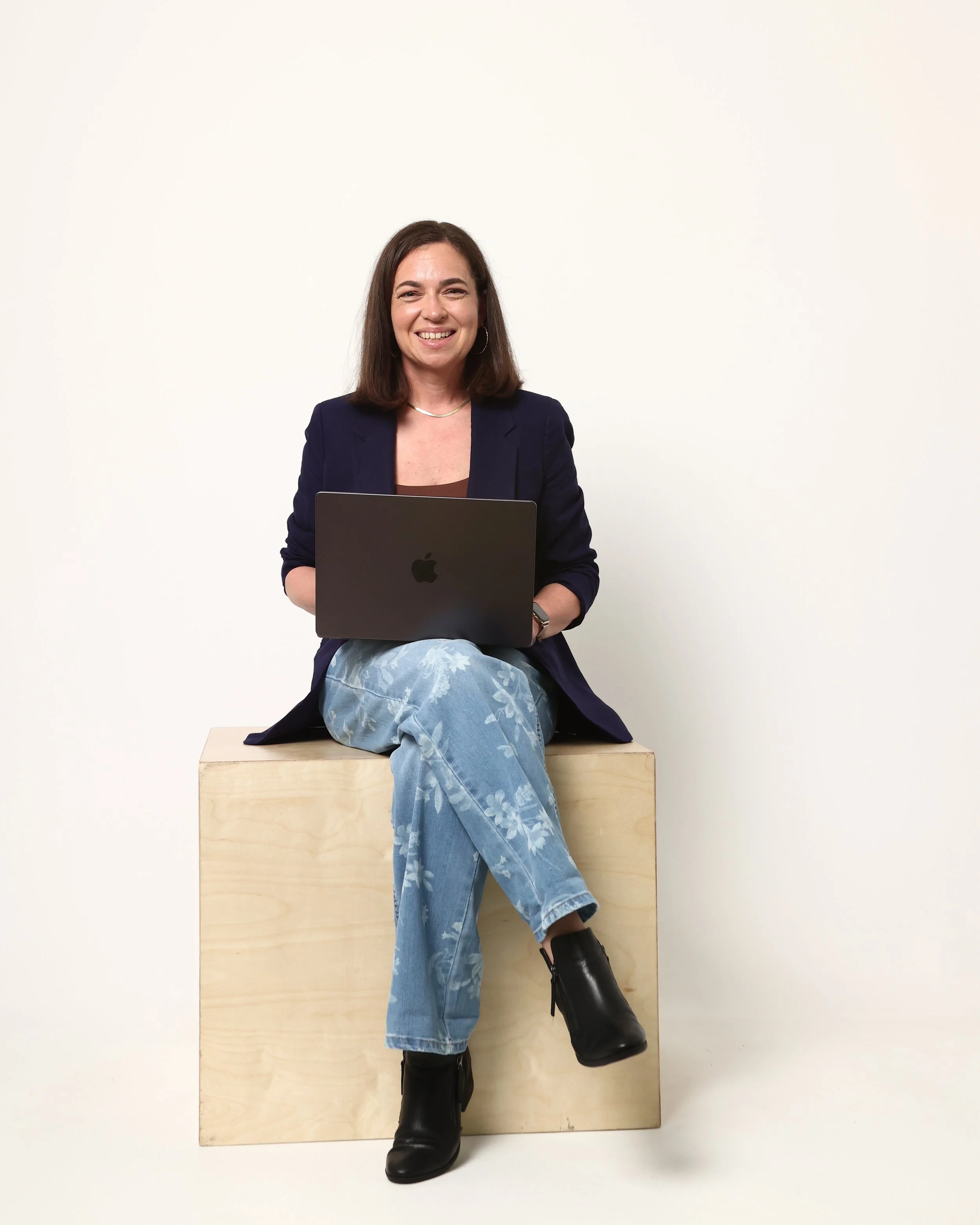 Lindsay smiling while sitting on a light wooden block, holding a silver laptop, against a plain light background. She wears a dark blazer, patterned light blue jeans, and black ankle boots.