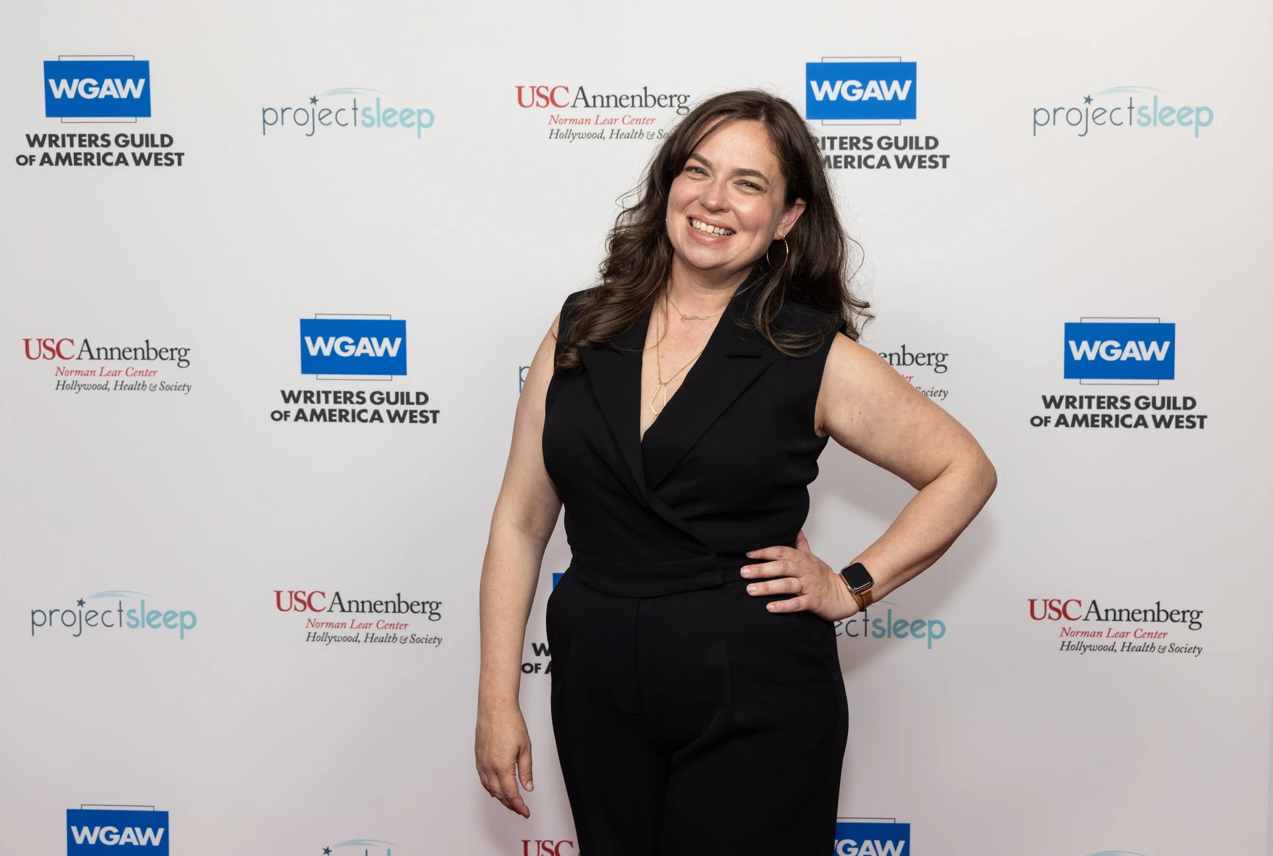 Lindsay smiling, wearing a black sleeveless jumpsuit, gold jewelry, and a watch, posing in front of a backdrop with logos for the Writer's Guild, Project Sleep, and USC Annenberg Hollywood Health and Society.