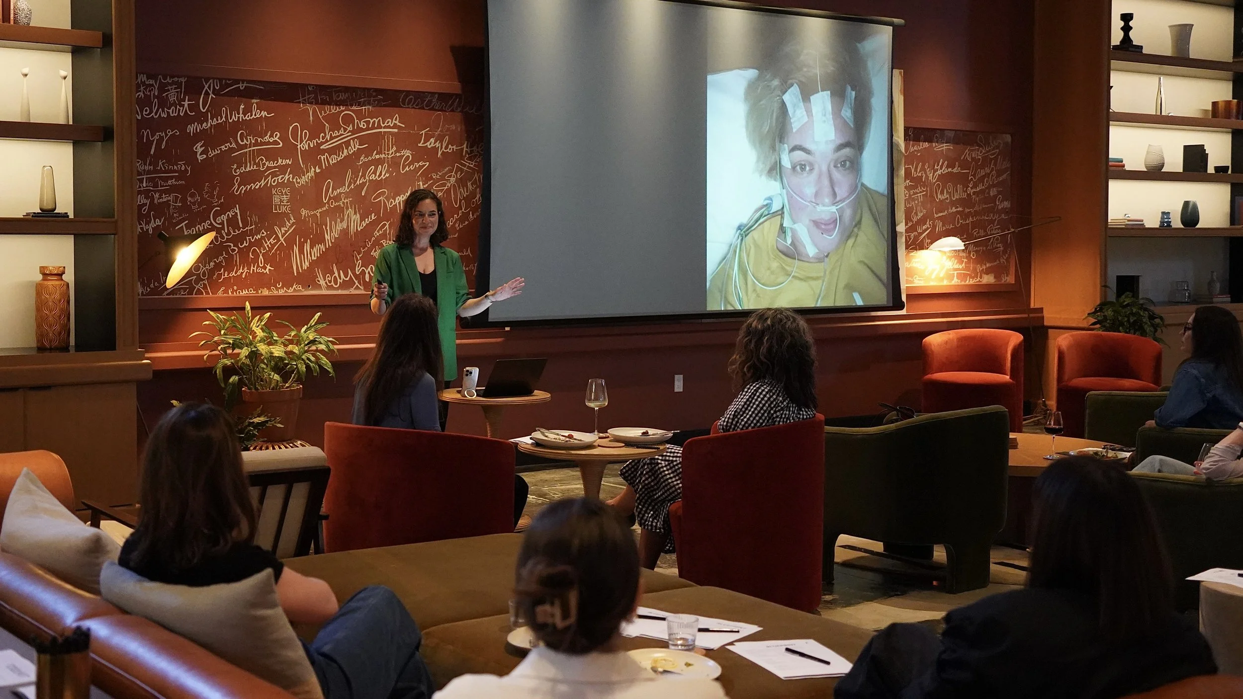 Lindsay in a green blazer giving speaking at Chief, with a large screen displaying a picture of Lindsay during a sleep study. Audience members are seated, some with drinks and notepads.