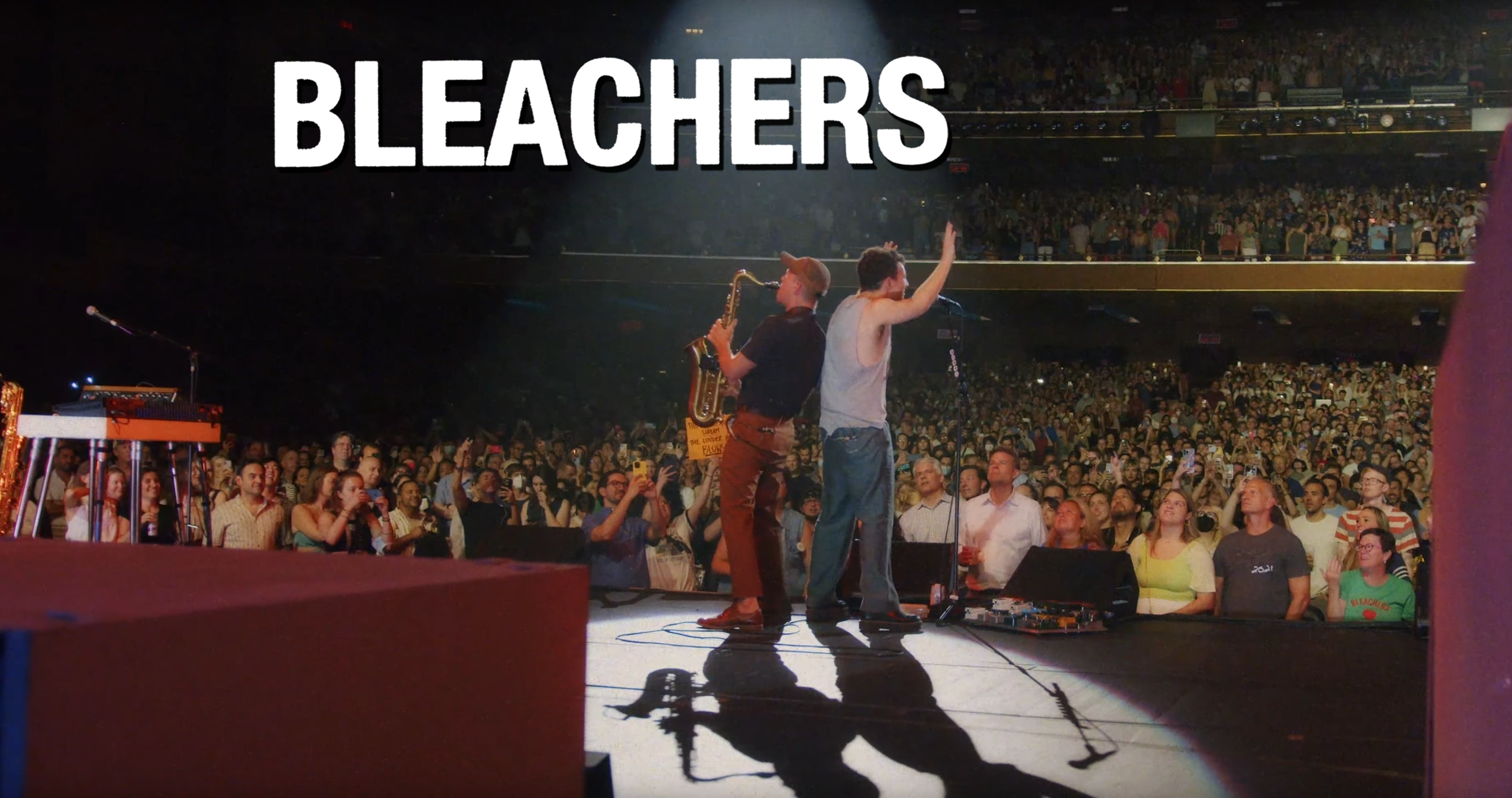 Two musicians performing on stage in front of a large audience at a concert. One is playing a saxophone, and the other appears to be singing or speaking into a microphone. The word 'BLEACHERS' is displayed in large white letters at the top of the ima