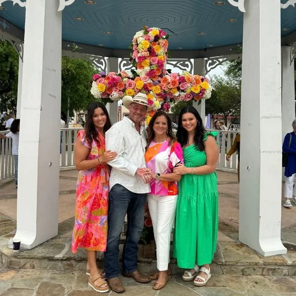 Four people pose in front of a flower-covered cross in an outdoor pavilion.