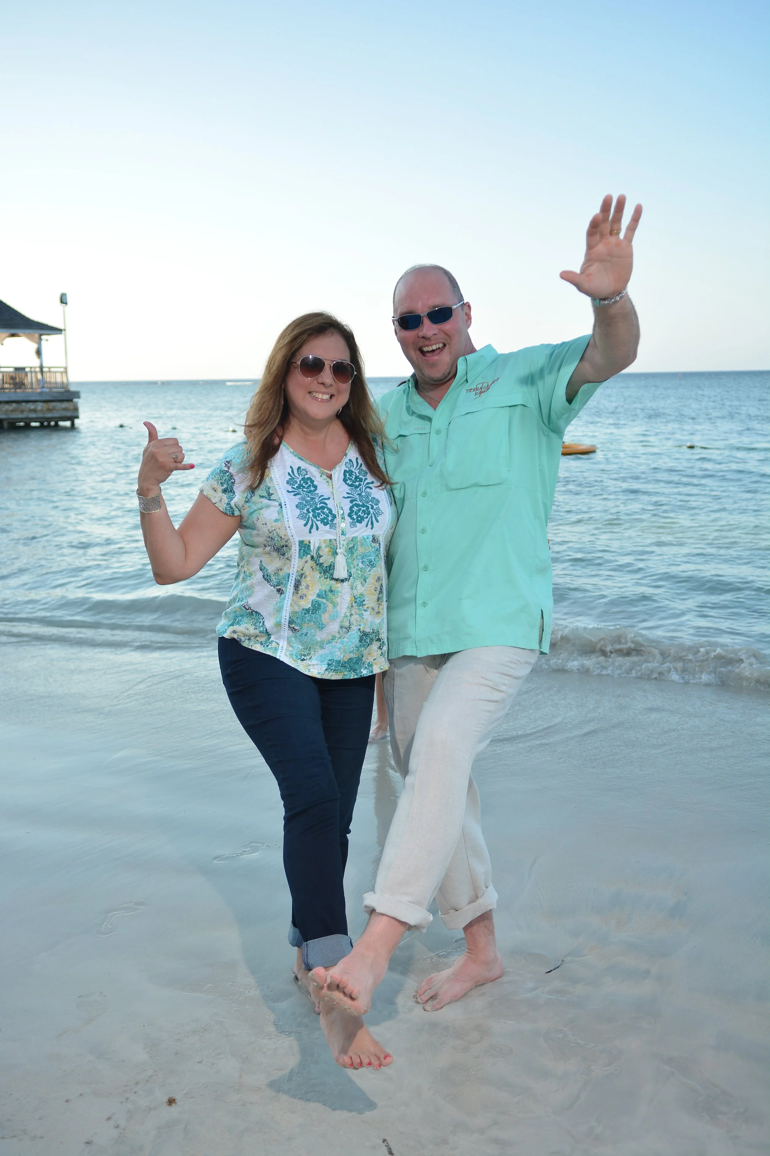 A happy couple standing on the beach in front of the ocean. They are smiling and waving, dressed in casual beach attire, with the woman pointing at the man. The sky is clear, and there's a small pier with a gazebo in the background.