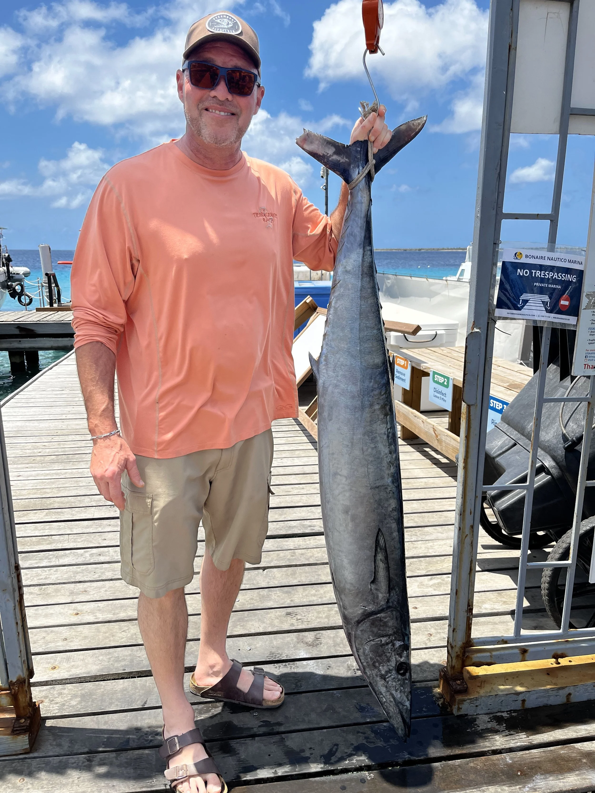 Man wearing sunglasses, a cap, and an orange shirt holding a large fish on a dock by the ocean.