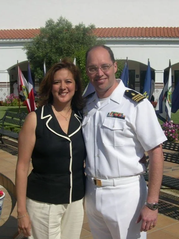 Man in a white military uniform standing next to a woman in a black sleeveless top outdoors with Flags and benches in the background.