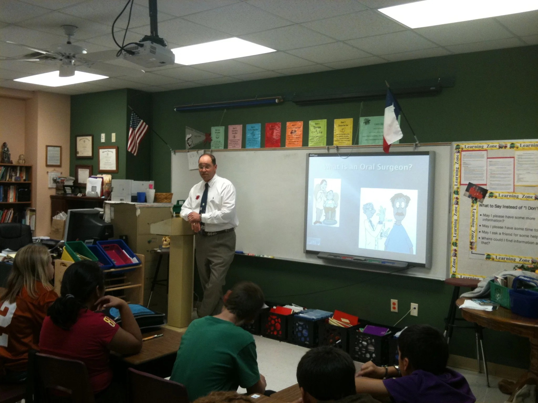A male teacher in a white shirt and gray tie stands at the front of a classroom, teaching students seated at desks. The classroom has a green wall, with a whiteboard and a projection screen displaying a slide about oral surgeons. The American and Fre