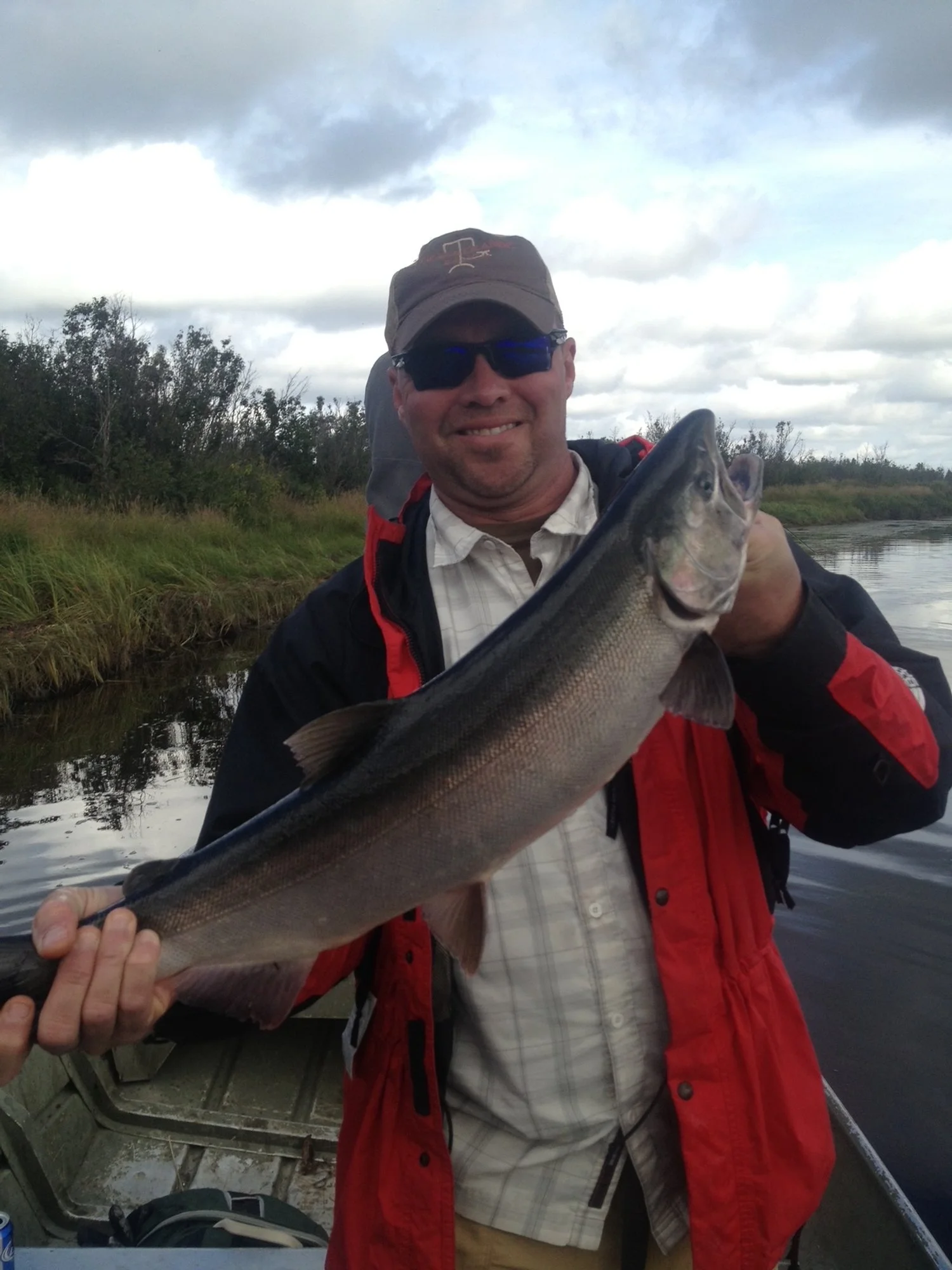 A man wearing sunglasses, a baseball cap, a plaid shirt, and a red jacket holding a large fish on a boat in a rural waterway with trees and cloudy sky in the background.