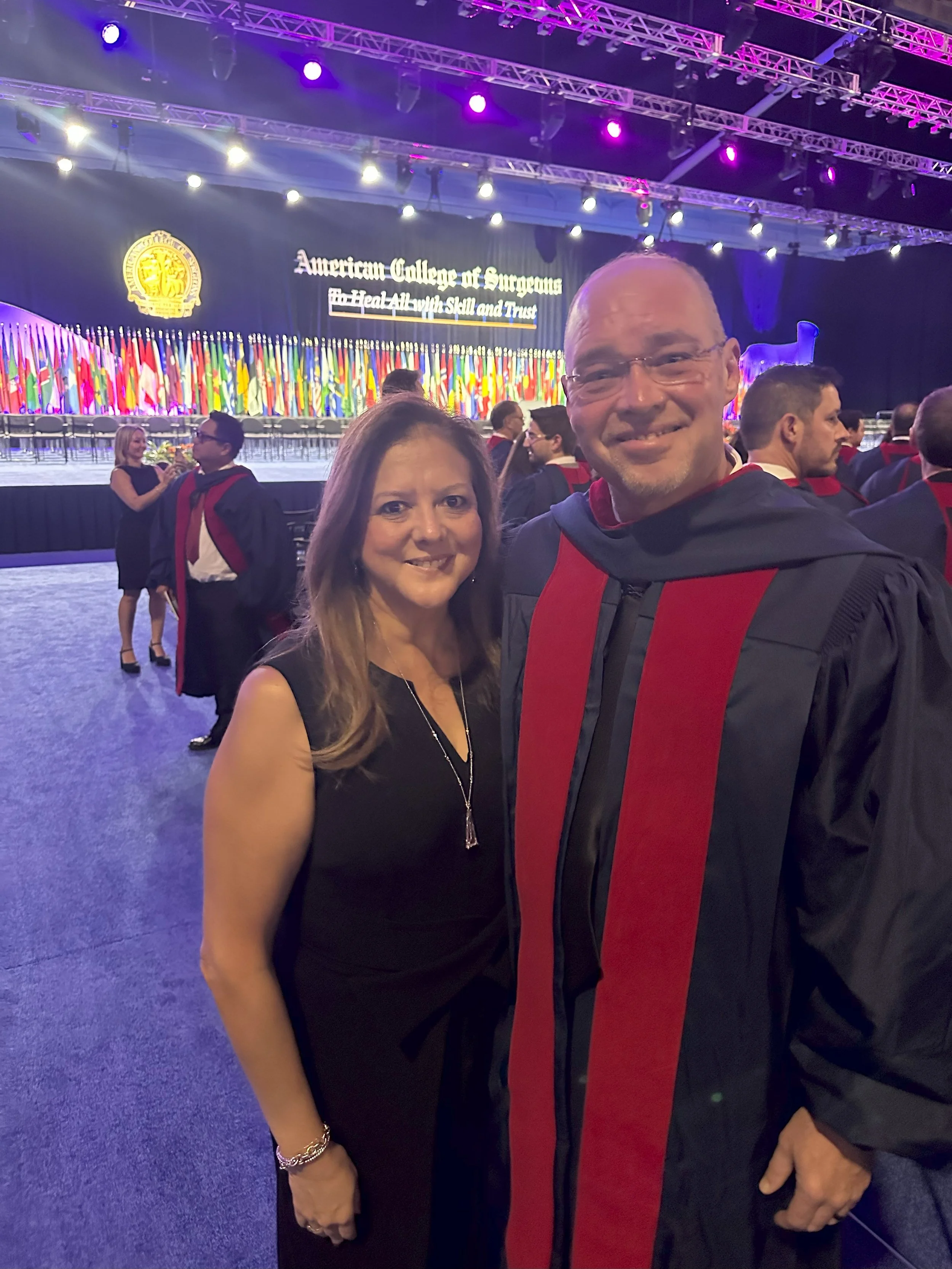 A woman and a man in academic regalia at a graduation ceremony with many national flags and a large banner reading 'American College of Surgeons' in the background.