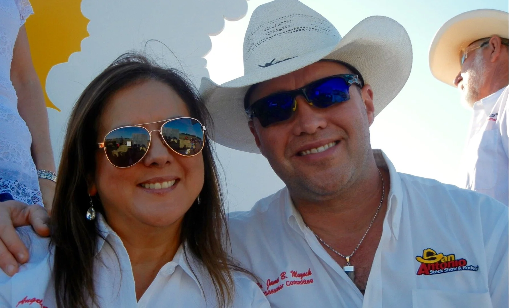 A smiling woman wearing sunglasses and earrings, and a man smiling wearing sunglasses, a cowboy hat, and a white shirt with the logo 'San Antonio Stock Show & Rodeo'.