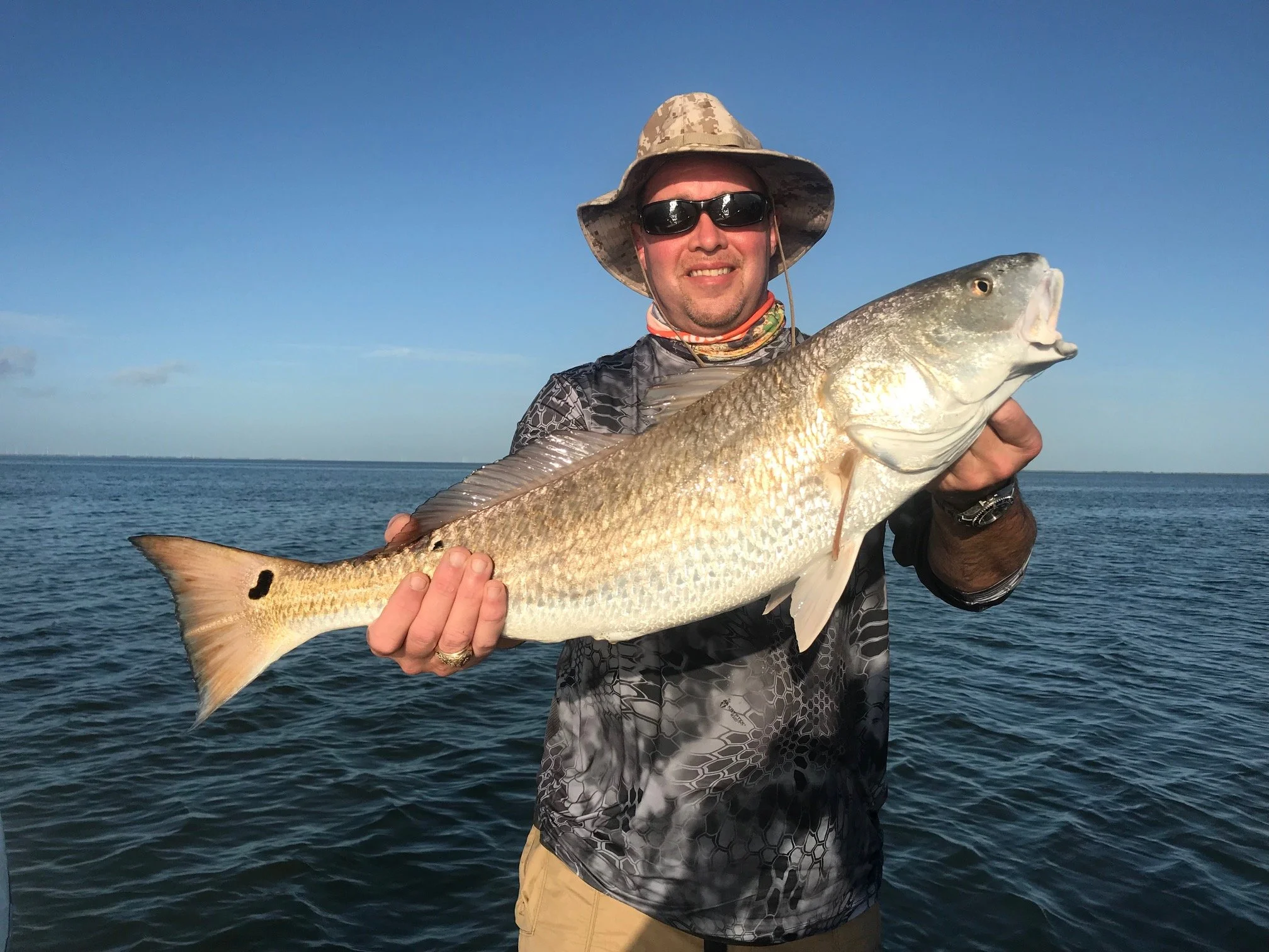 Man wearing hat and sunglasses holding a large fish over a body of water