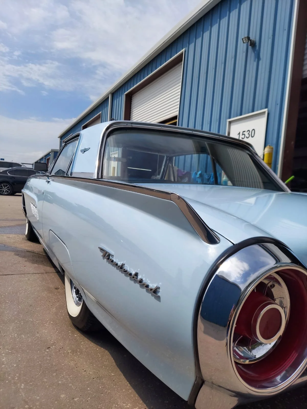 A vintage blue and white Thunderbird car parked outside a blue industrial building with a roller shutter door.