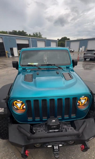 Front view of a teal Jeep Wrangler with black grille and bumper, parked outside a warehouse.