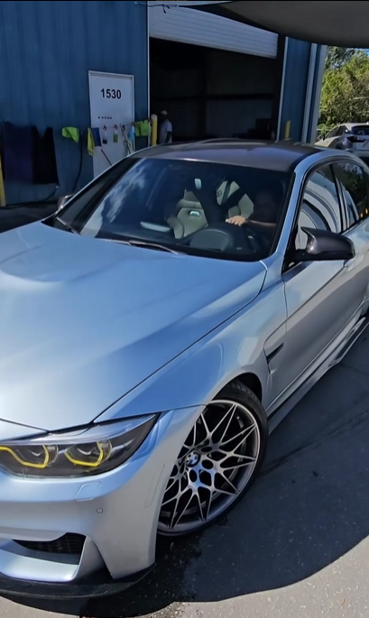 Silver BMW car parked near a building with a person inside, and trees in the background.