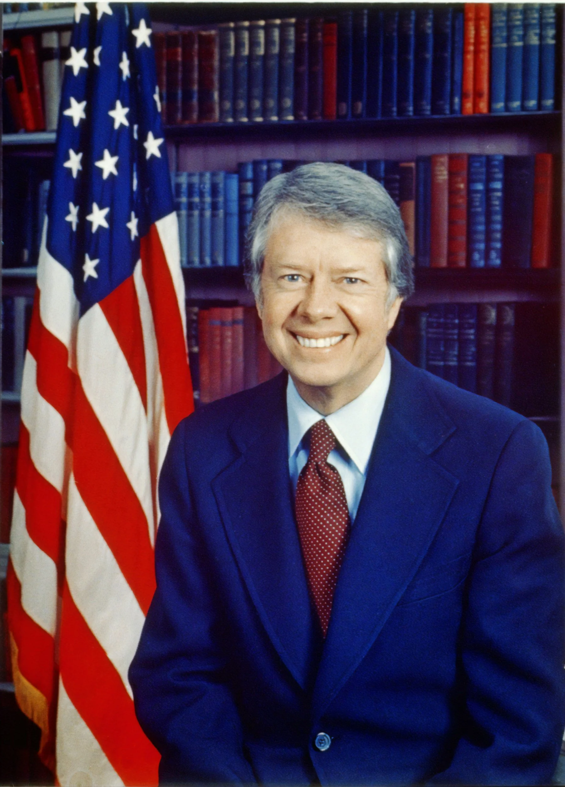 A man in a navy suit with a red tie sitting in front of an American flag and a backdrop of a bookshelf with red and blue books.