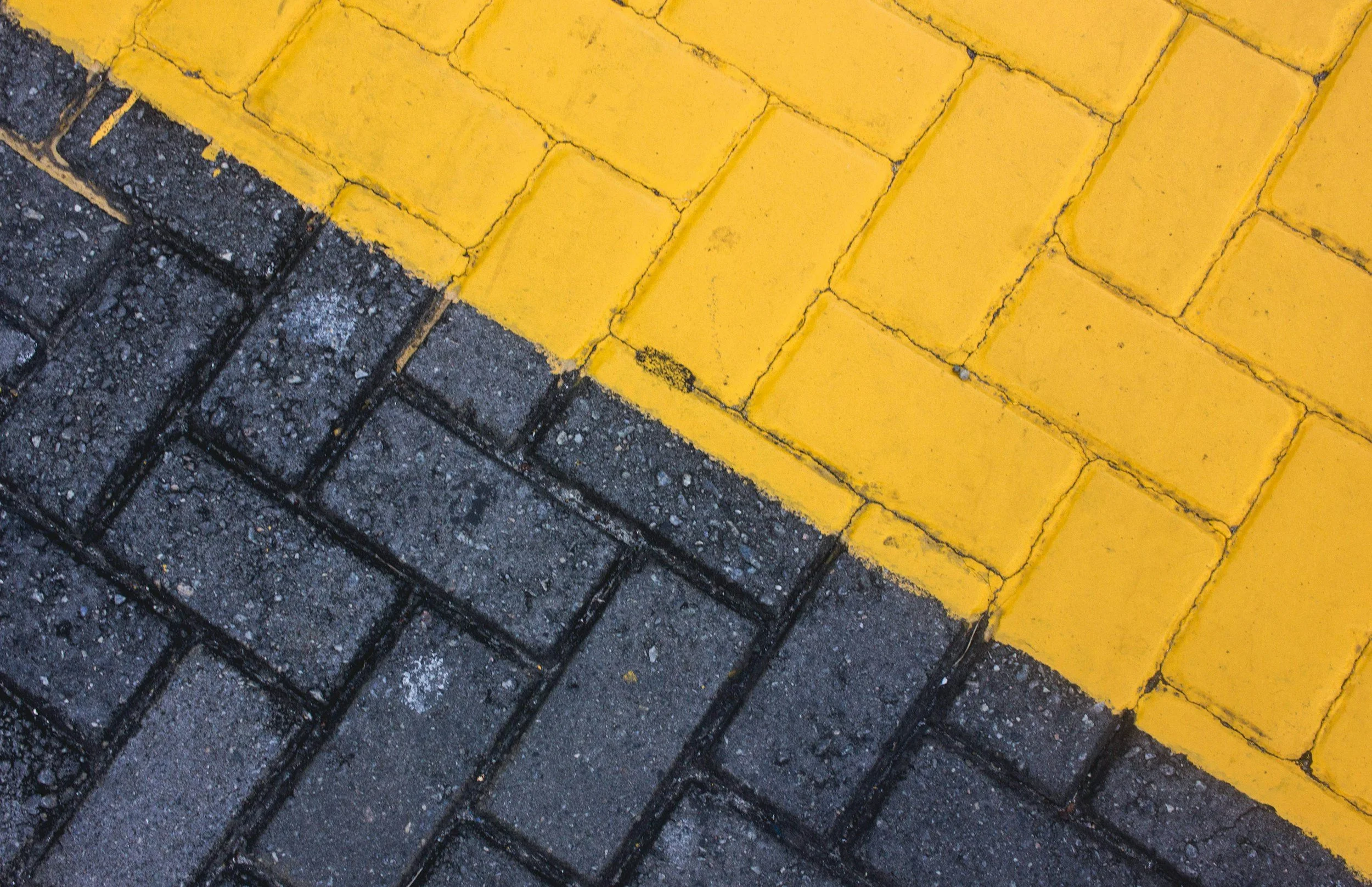Close-up of a street corner showing a yellow painted curb next to gray asphalt paving with brick-like pattern.