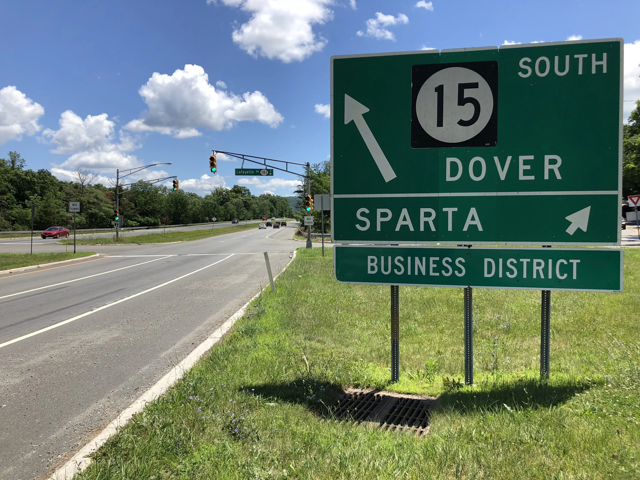 Green highway directional sign indicating route 15 south to Dover and Sparta, with a business district exit to the right, located by a road with cars and traffic lights in the background under a partly cloudy sky.