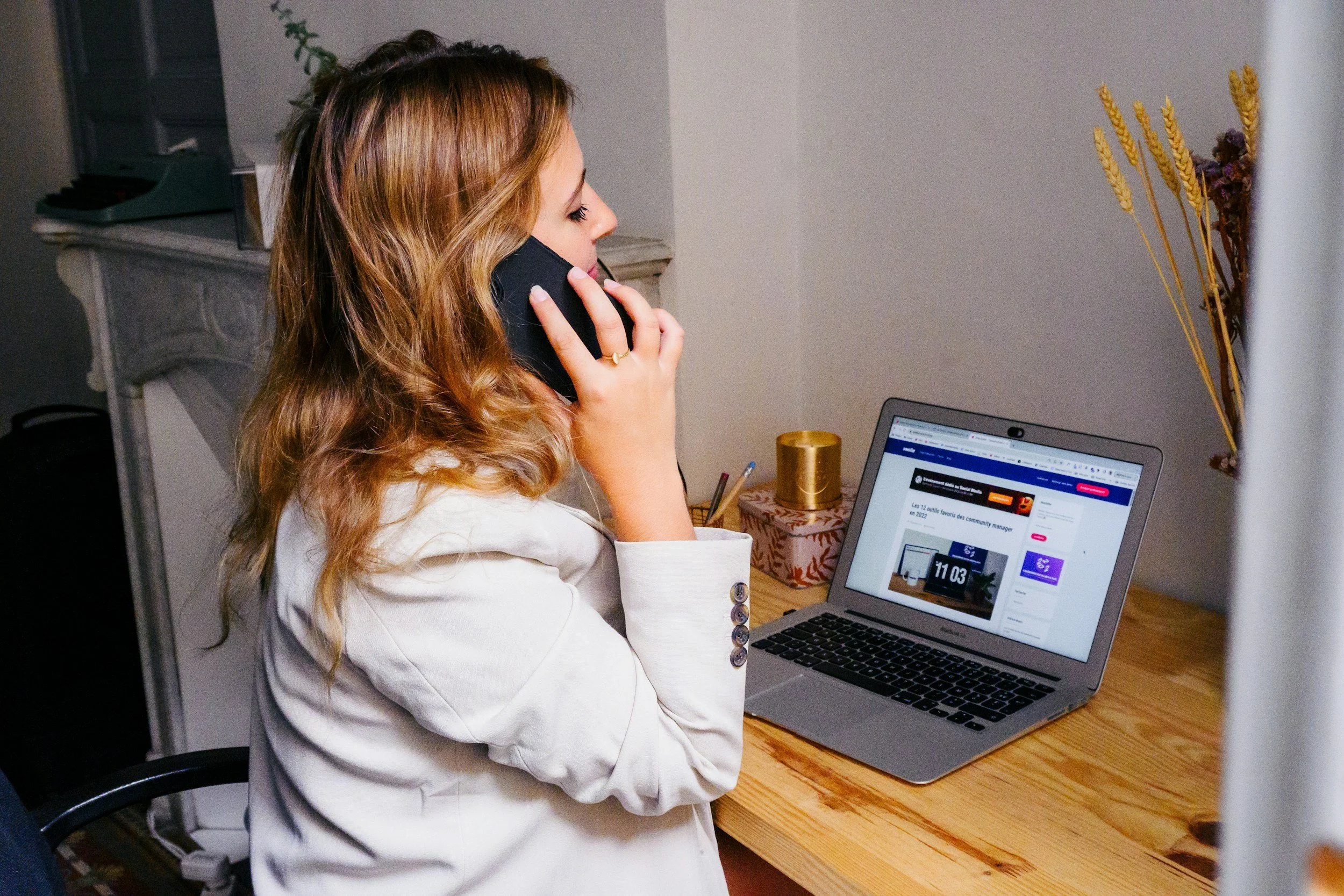 Woman with brown hair and white blazer talking on a cordless phone at a wooden desk, with a laptop open to a webpage, a gold cup, and dried flowers in the background.