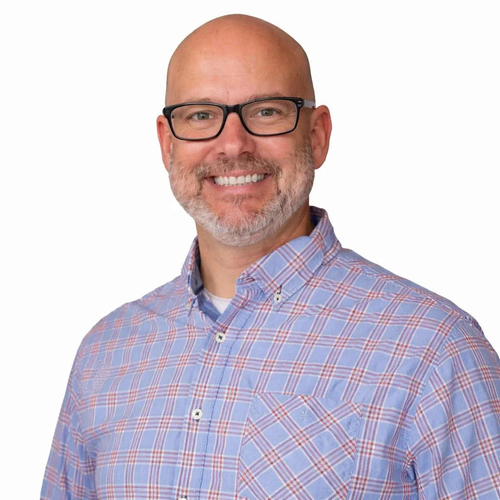 A smiling middle-aged man with a bald head, glasses, and a beard, wearing a blue and red plaid button-up shirt, against a plain white background.
