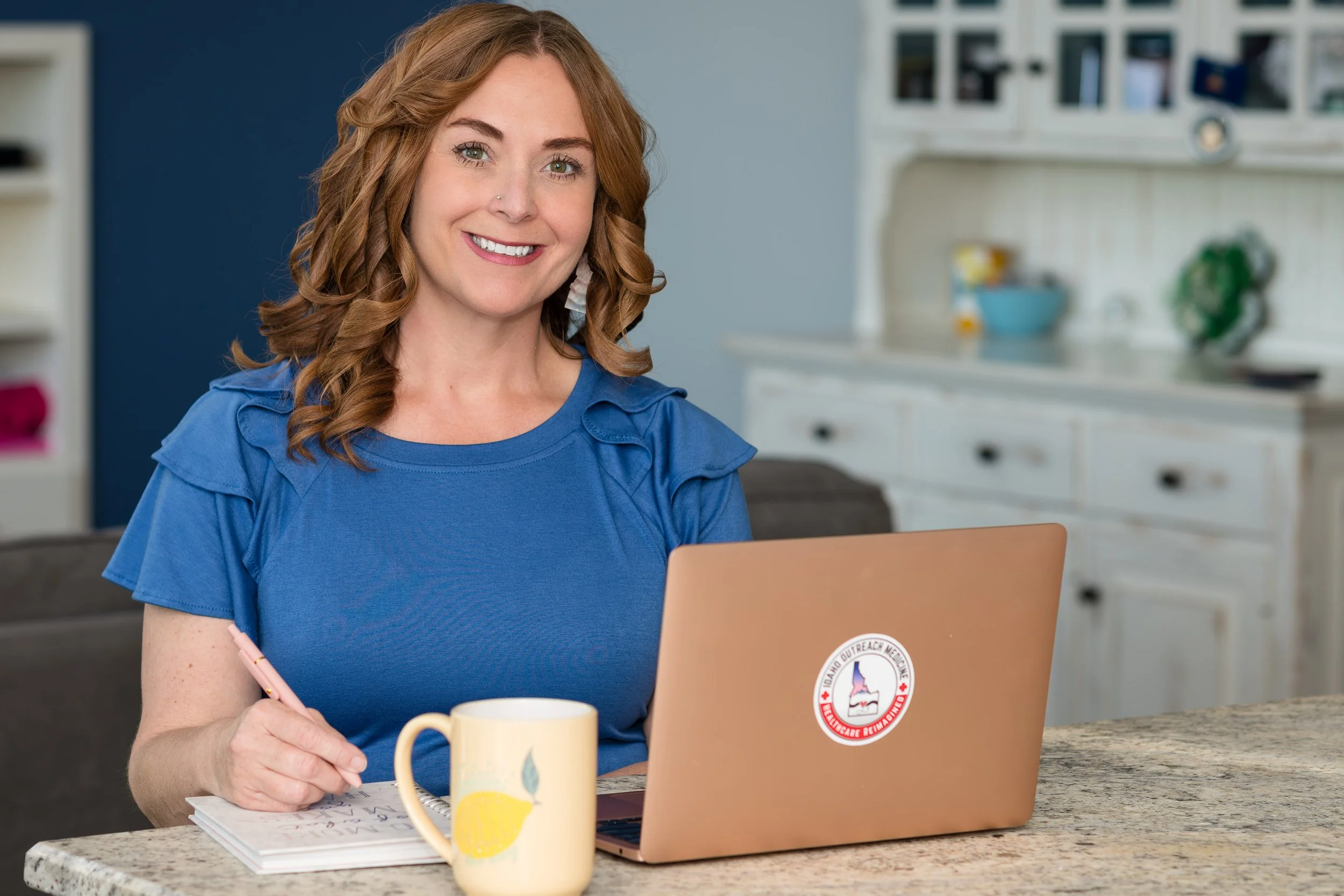 A woman with wavy red hair in a blue top sitting at a kitchen counter with a laptop, a notebook, a pen, and a yellow mug with lemon and leaf illustrations, smiling at the camera.