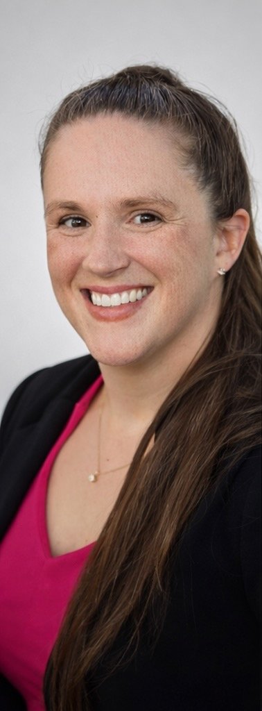 A woman with long brown hair, smiling, wearing a black blazer and a pink top, standing against a plain light gray background.