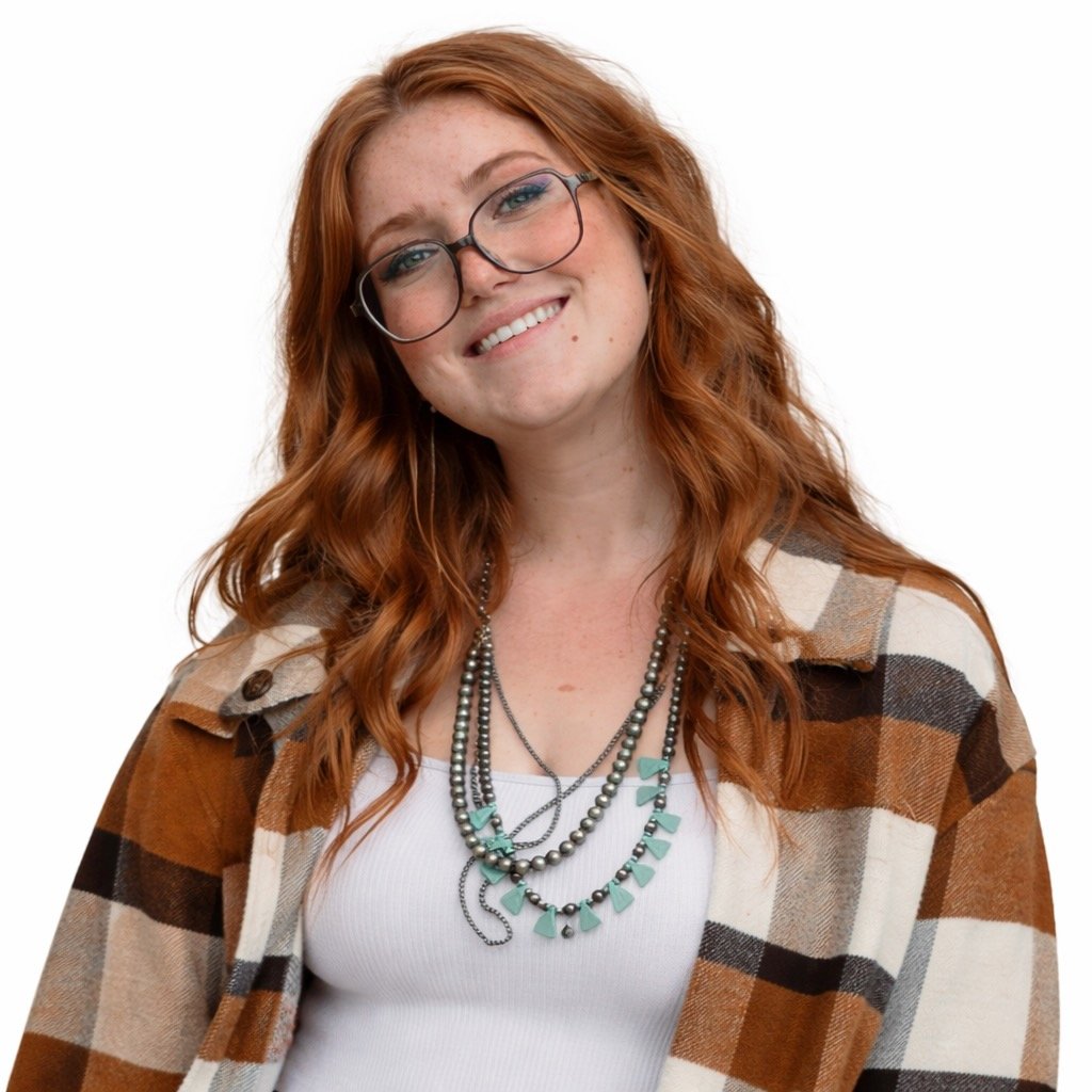 A young woman with long red wavy hair, wearing glasses, a white tank top, layered necklaces, and a brown and beige plaid shirt, smiling at the camera against a plain white background.