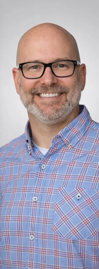 A smiling man with a bald head, glasses, and a gray beard, wearing a blue plaid button-up shirt.
