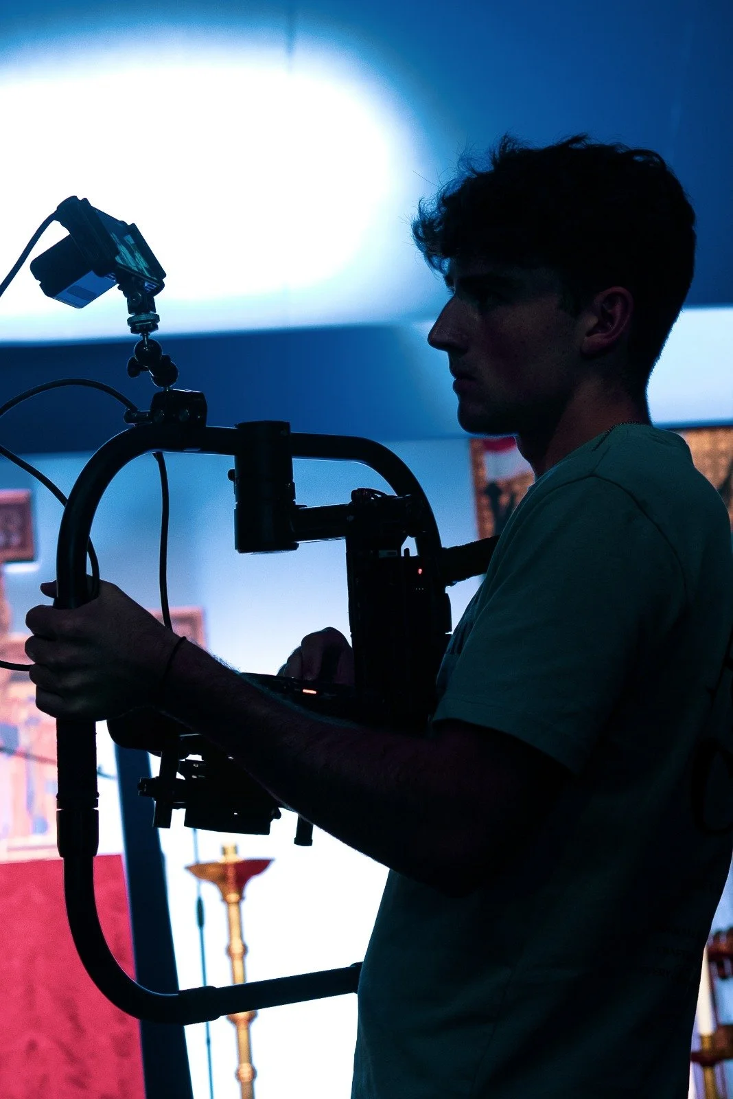 Silhouette of a videographer or photographer operating camera equipment indoors, with background including artwork and candle holder.