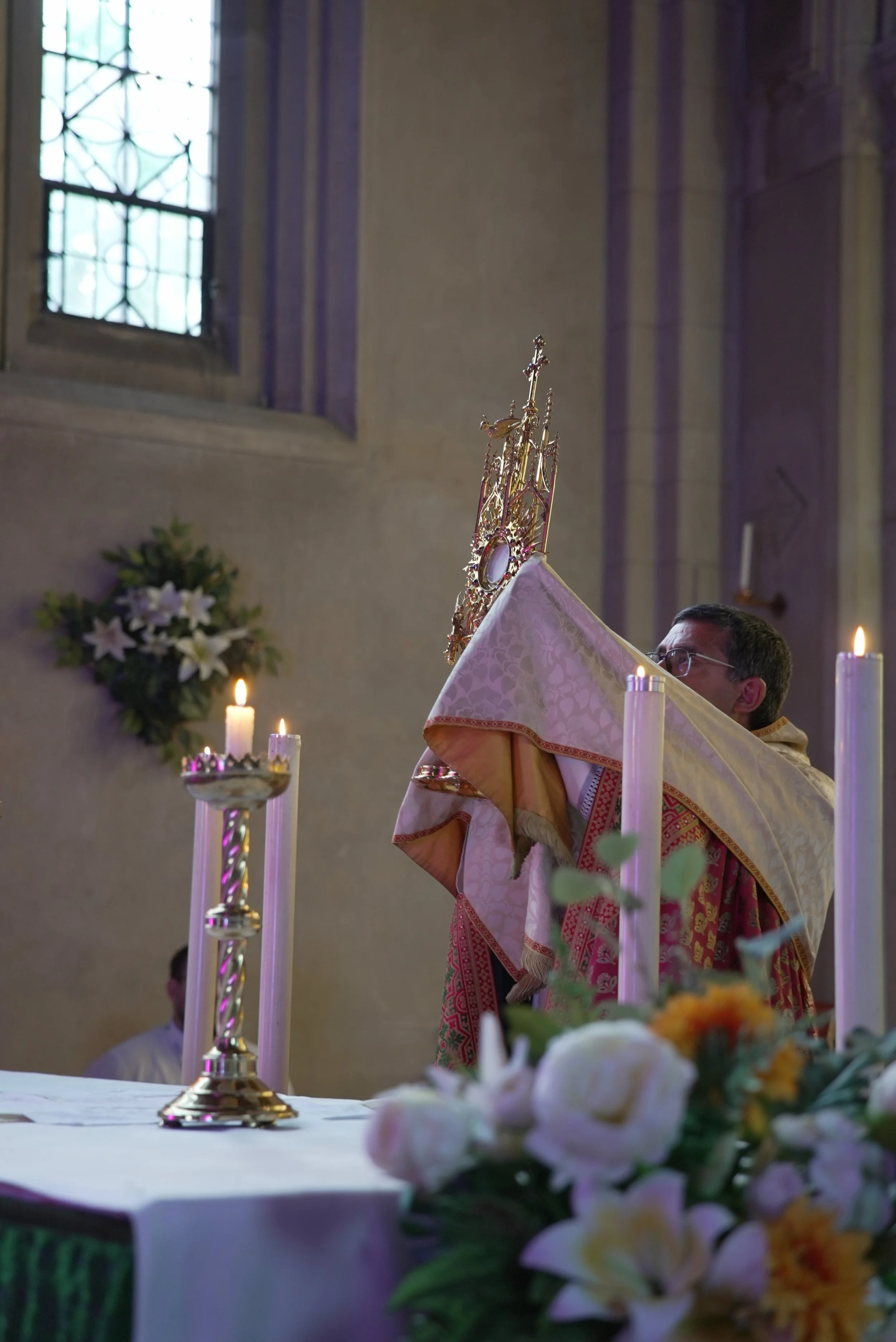 A priest stands behind an altar during a religious ceremony, holding a gold and white cloth with an ornate gold crown or monstrance on top. The altar is decorated with white and pink flowers and tall candles, and there is a stained glass window in the background.