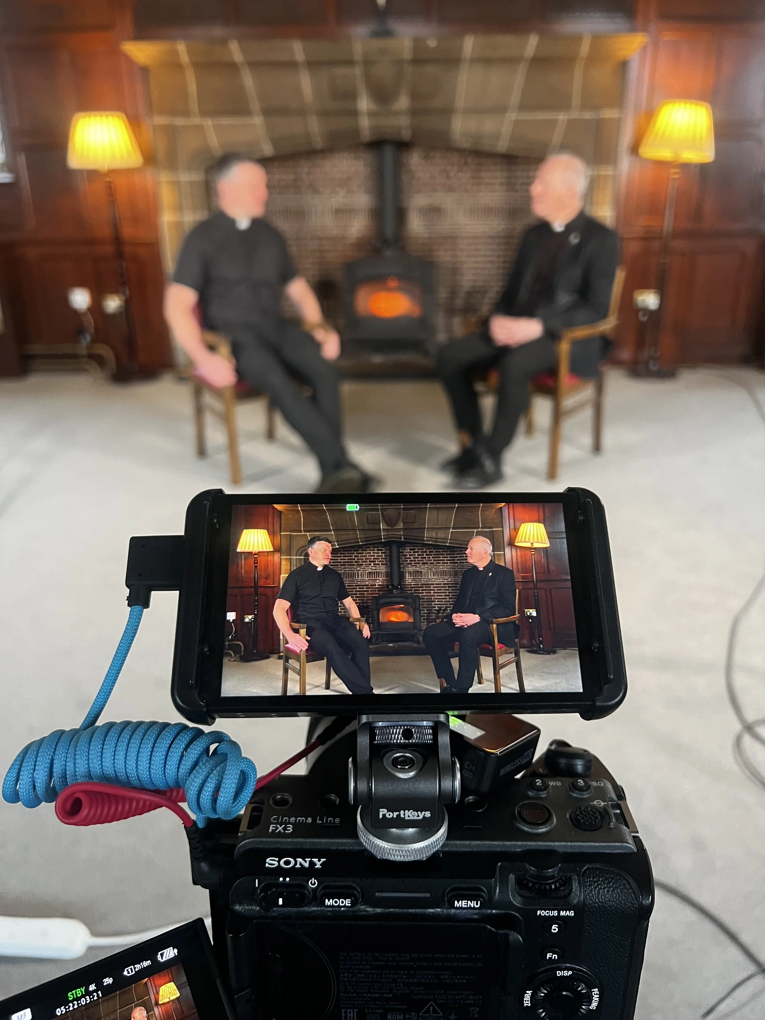 A video camera facing two men seated in front of a brick fireplace, engaged in a conversation. The room has wooden paneling and two yellow-lit lamps on either side of the fireplace.