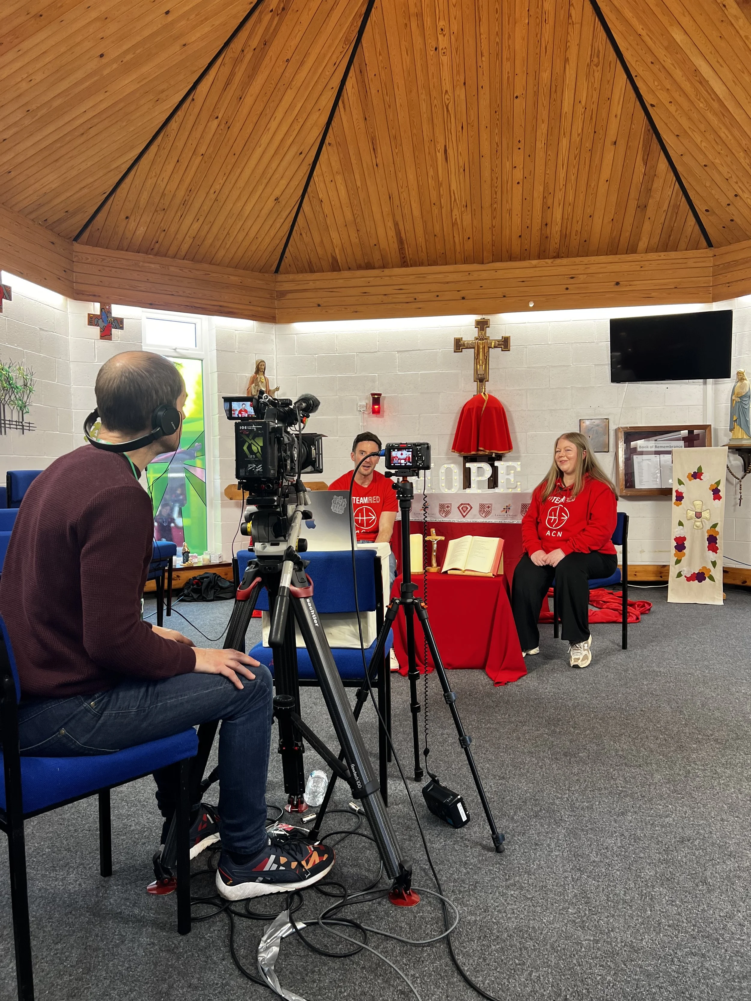 Two people seated at a table with a religious background, being filmed by a cameraman. The setting includes a cross, a statue, and signs that say 'HOPE' and 'TEAM RED.'