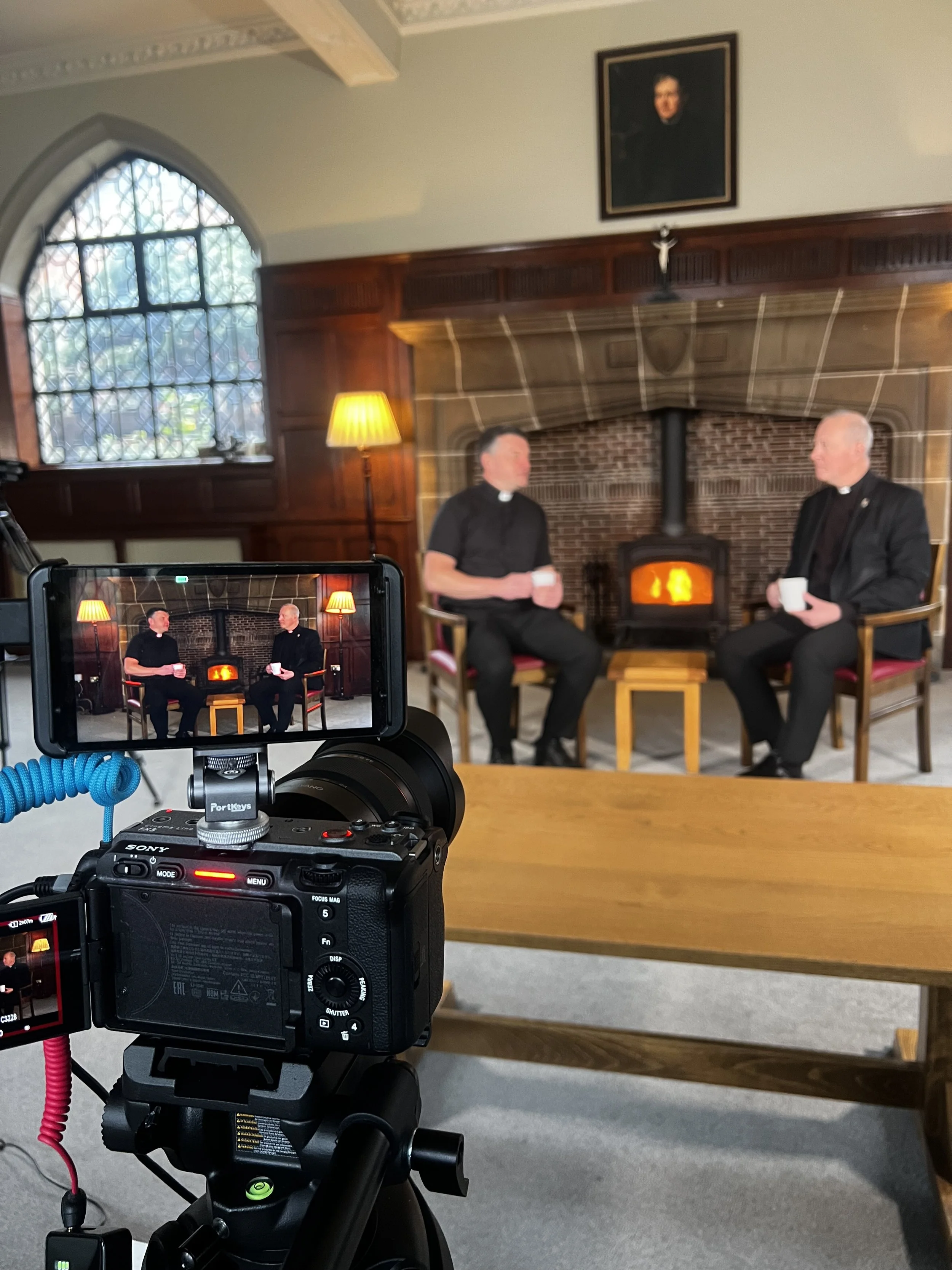 Two priests in black clerical shirts sitting in chairs near a fireplace, engaged in conversation while holding mugs, with a camera filming them in a room with wooden paneling, a large window, and religious artwork above the fireplace.