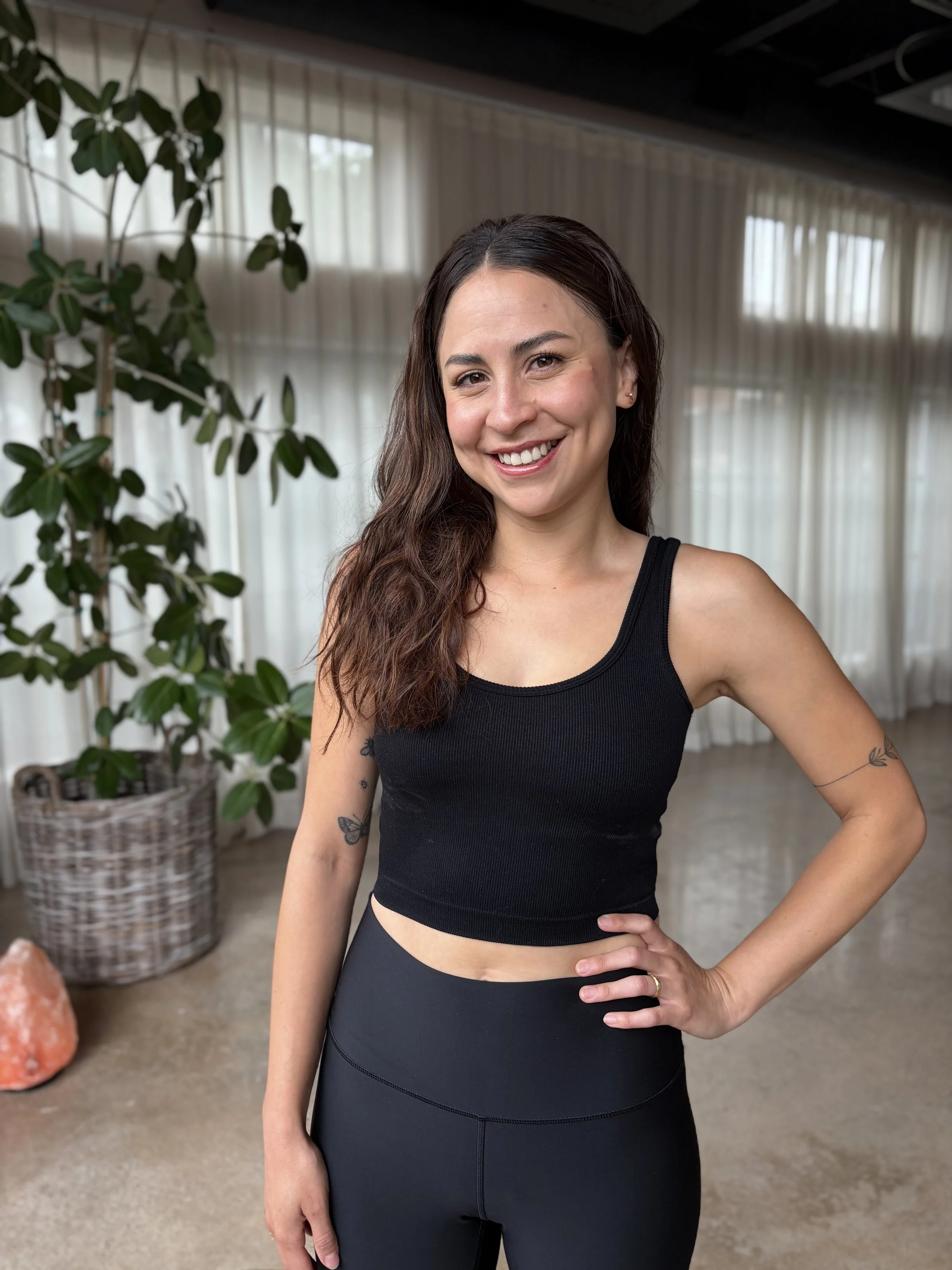 A woman with long wavy brown hair wearing a black sports bra and black leggings, standing inside a room with large windows and white curtains, with a plant and salt lamp in the background.