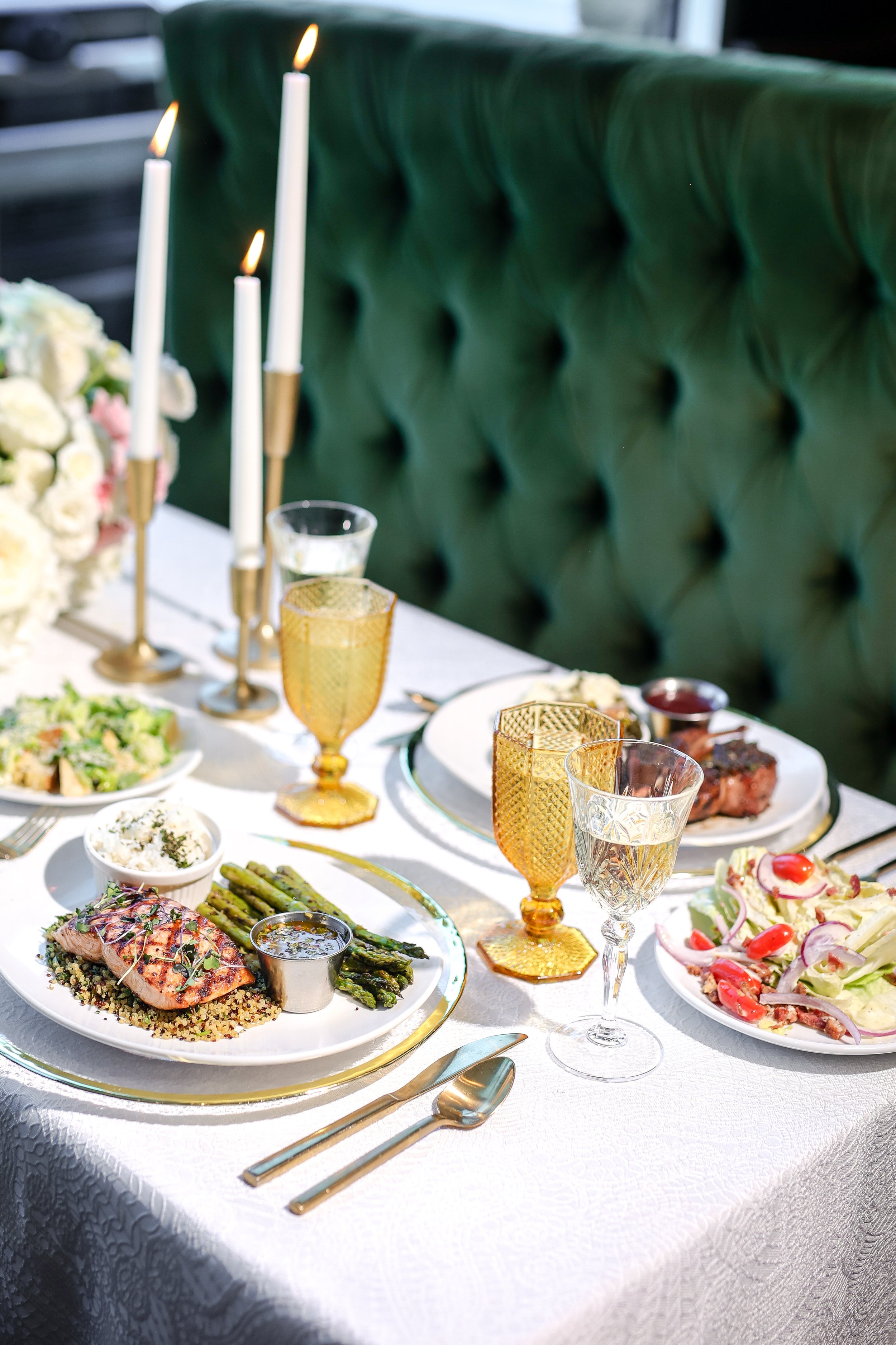 A beautifully set dining table with various dishes, gold-rimmed glasses, white candles, and floral decorations, likely for a special occasion or dinner event.