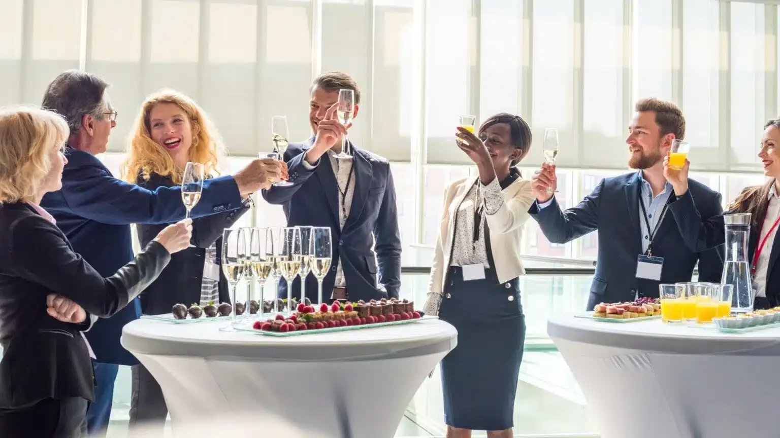 A group of professionals raising glasses in a toast at a corporate event with high tables and snacks.