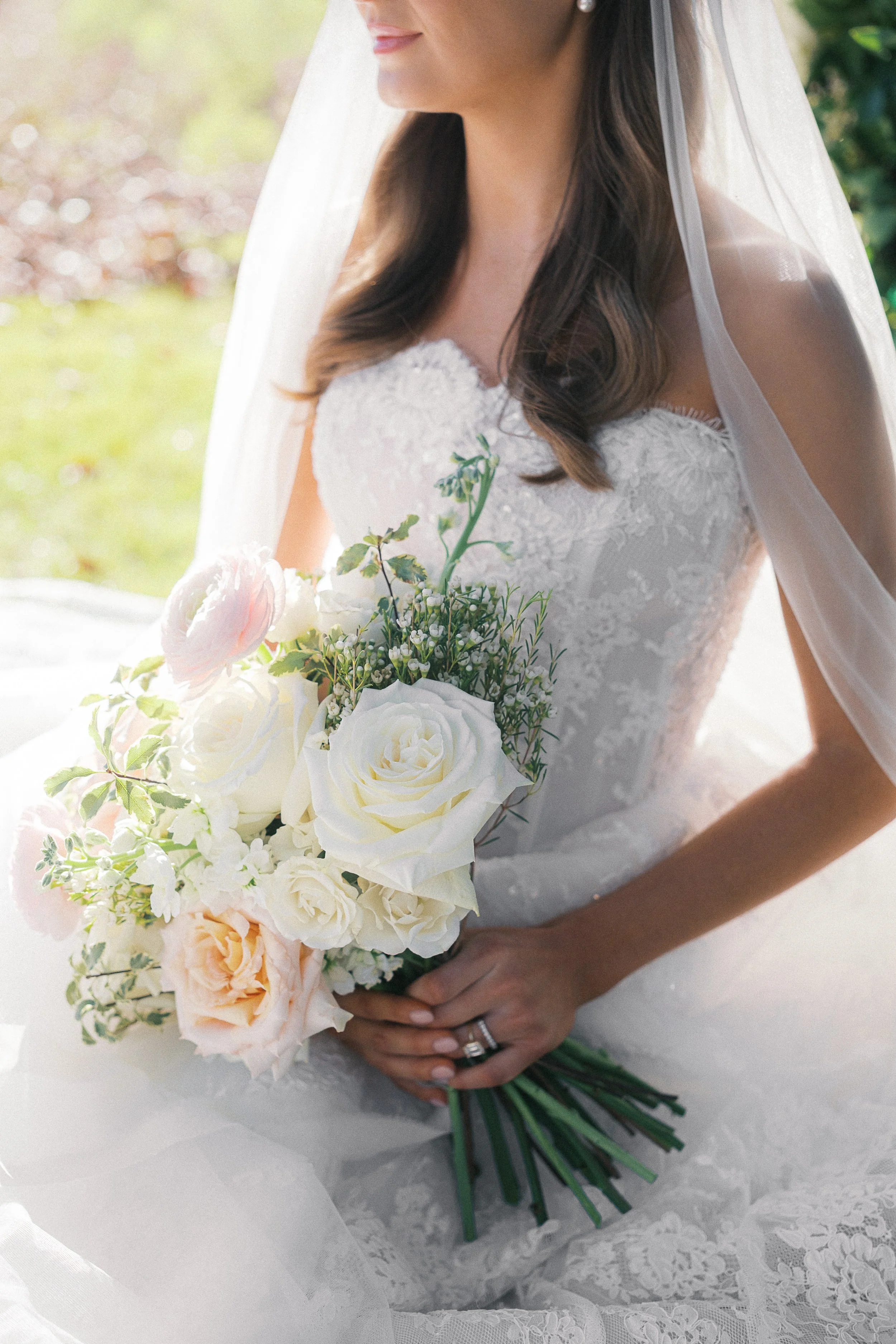 A bride in a lace wedding dress holding a bouquet of white and blush roses, greenery, and baby's breath, with a blurred outdoor background.