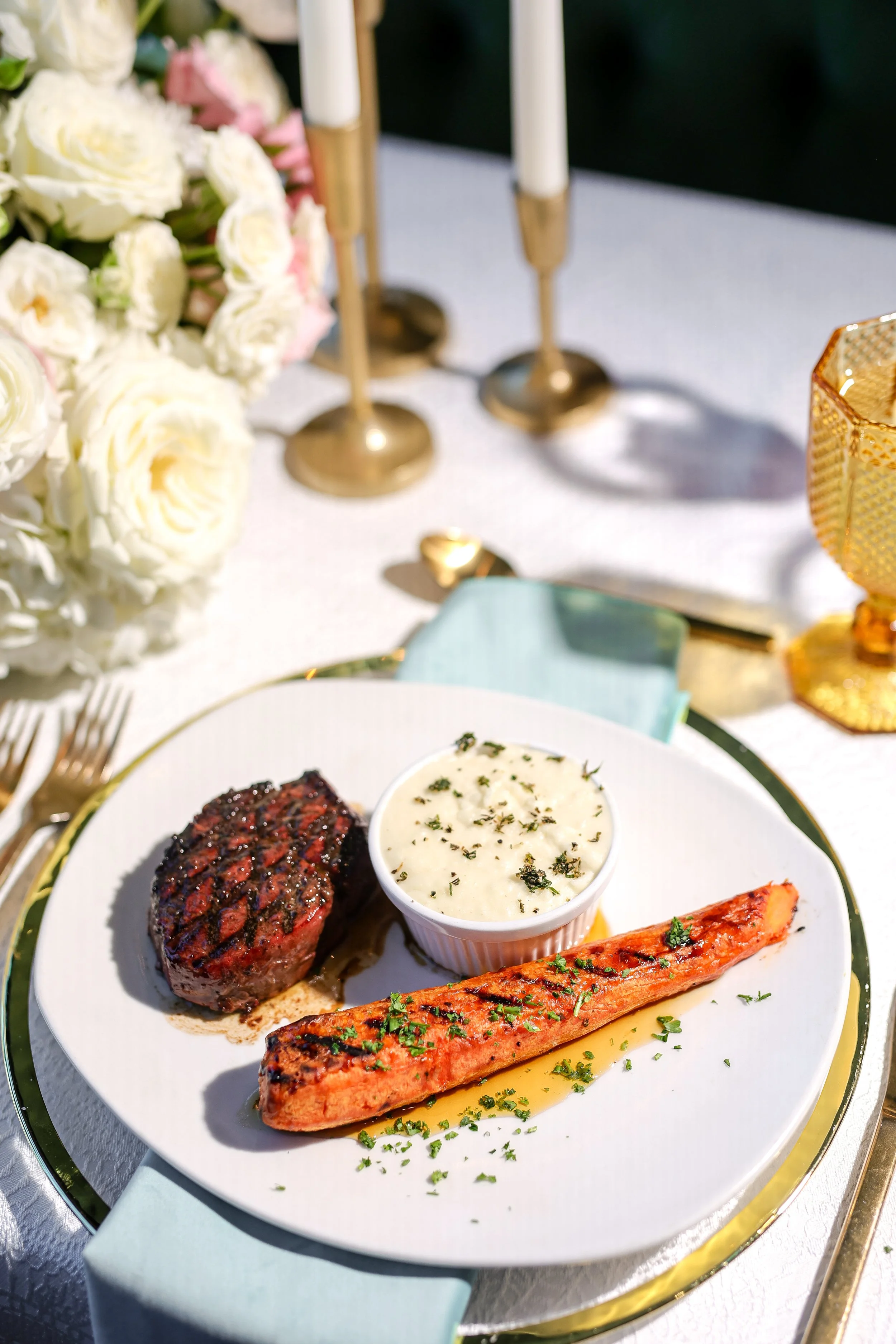 A plated meal with grilled steak, salmon, and a cup of mashed potatoes garnished with herbs, on a decorated table with candles, flowers, and gold accents.