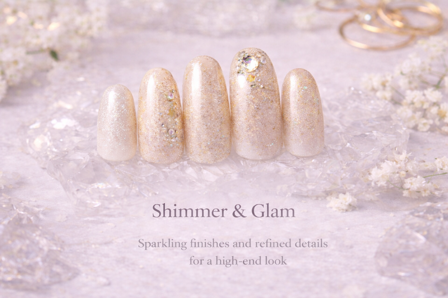 Close-up of five artificial nails with shimmering, glittery gold and white designs, decorated with rhinestones, on a reflective surface surrounded by small white flowers and golden rings.